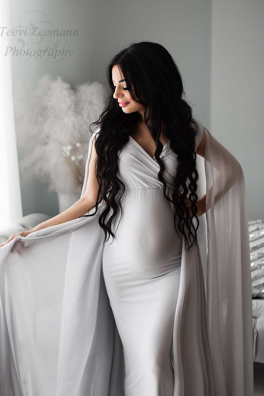 A pregnant woman is wearing a coolgrey dress. The dress is a tight fit model with loose chiffon grey fabric.