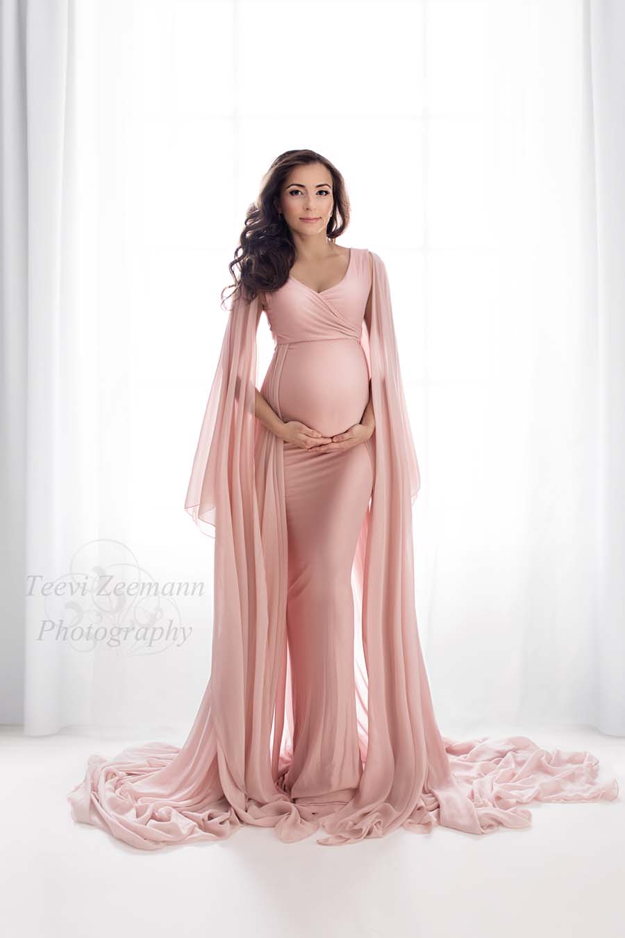 Brunette model looks at the camera while holding her bump with both hands. She is posing in a studio during a maternity photo shoot and wearing a dusty pink dress made of jersey and with long chiffon sleeves and train.