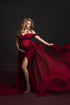 Blond pregnant model poses in a studio during a maternity photoshoot. She holds her bump with one hand while playing with the train from the dress with the other one. She wears a red dress with a side split and chiffon train.