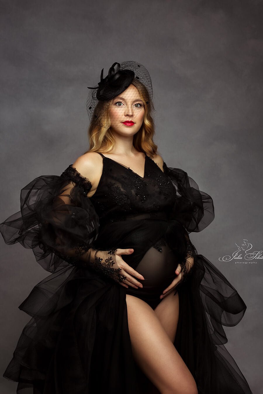 blond pregnant model wearing a black bodysuit made of mesh and bridal lace. she has a tulle cape to match the dramatic style.