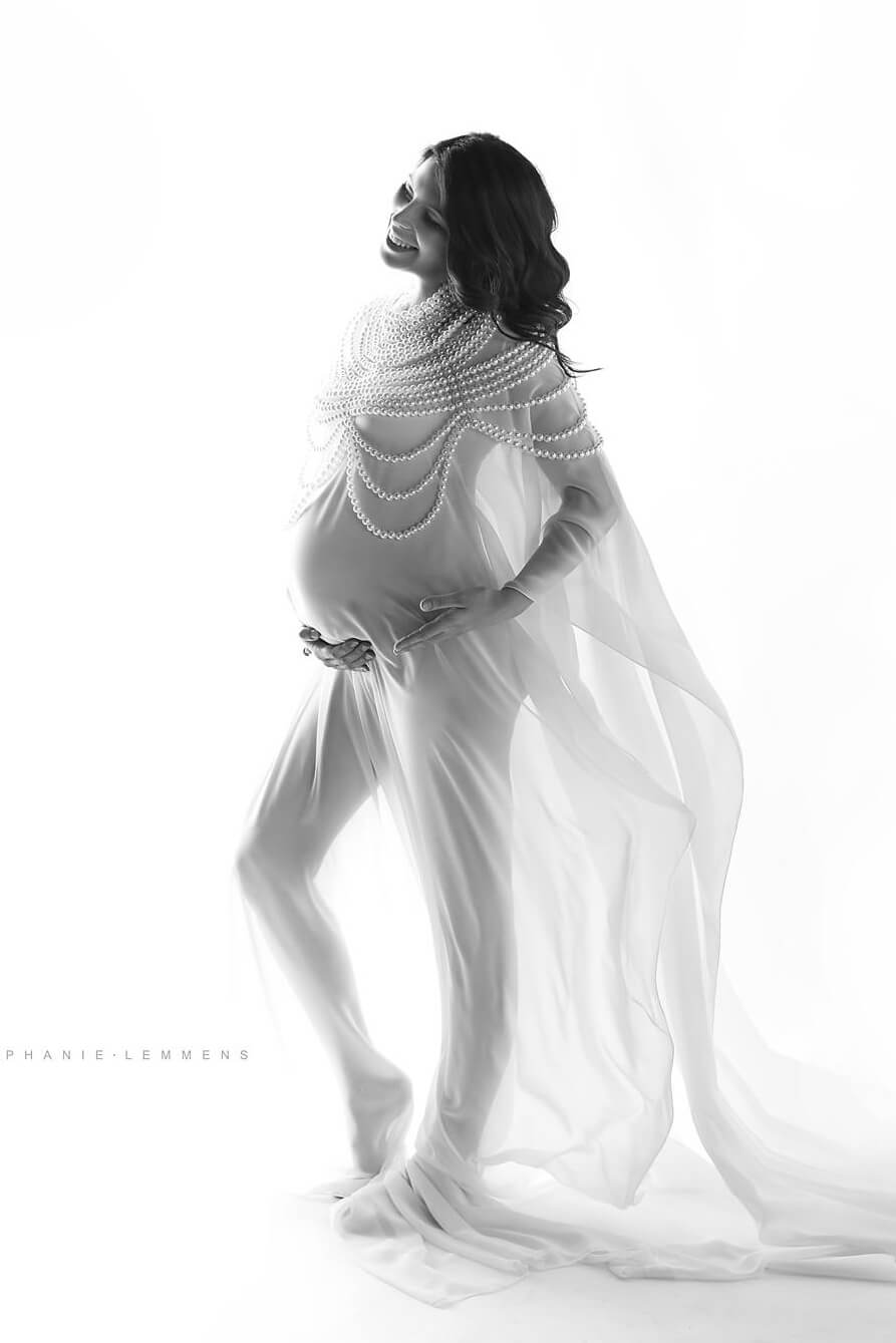 Pregnant model poses in a studio wearing a chiffon long cape in off white accessorized with a large pearl necklace. . She has long blond hair, has her eyes partially open while posing on her side and and holds her bump with both hands.