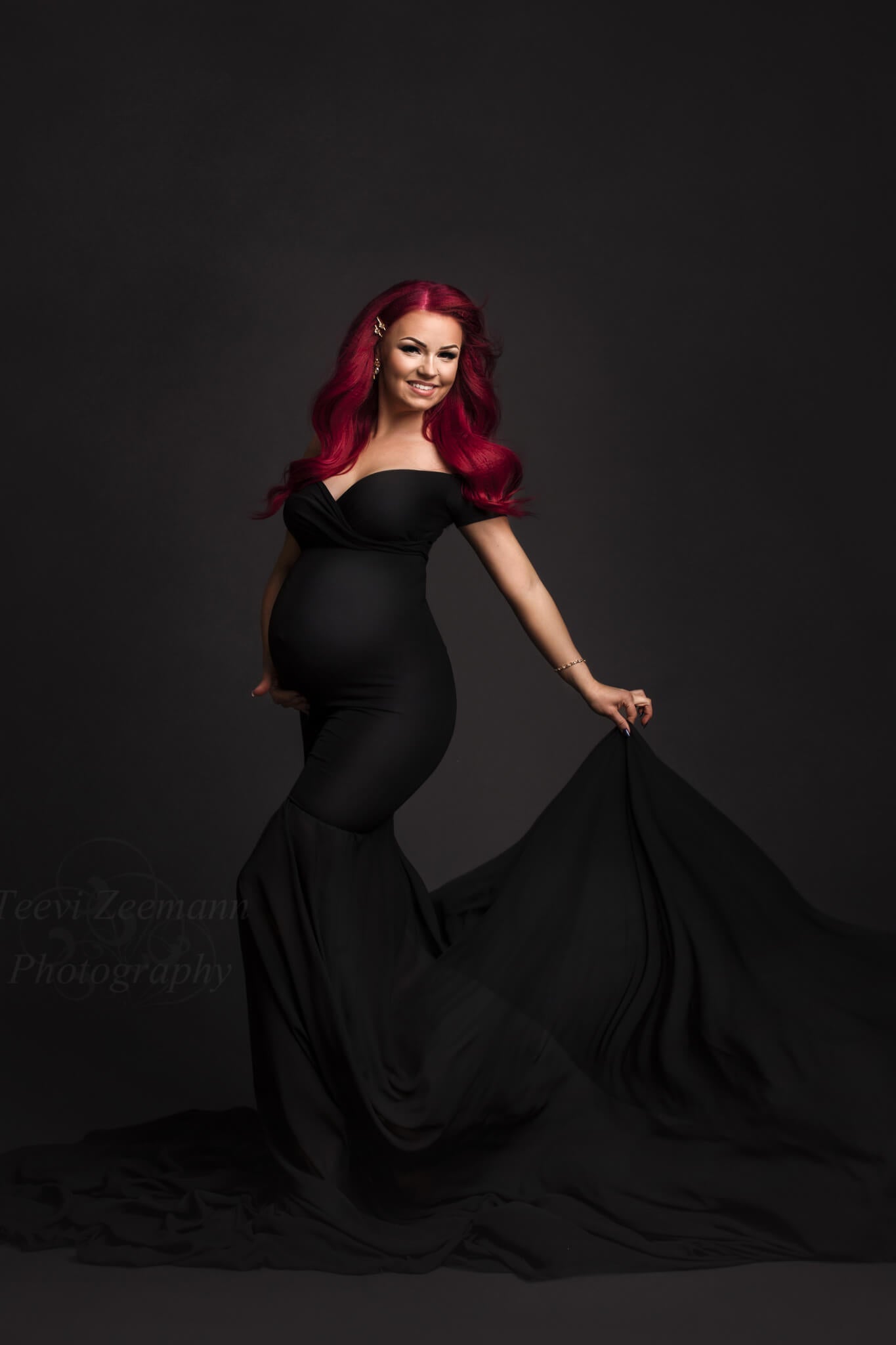 Pregnant model with bright, pink hair posing in a photography studio. She wears a black maternity dress with off shoulder sleeves and a mermaid skirt made of chiffon. The rest of the dress is made of jersey fabric which is tight fitting to her body. One hand is holding the skirt and the other one is under her belly. She is smiling and looking at the camera.