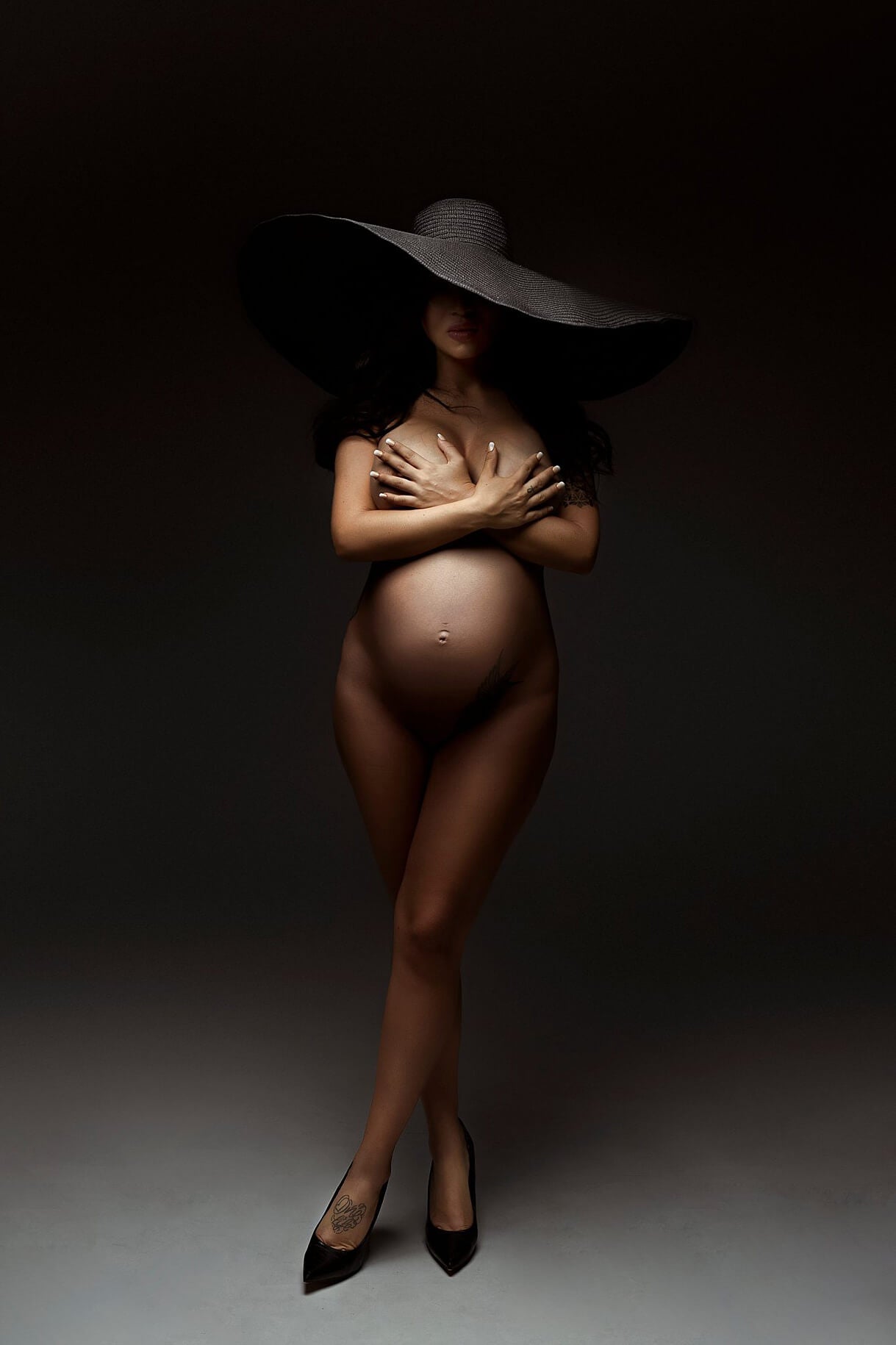 The woman is standing inside a studio. The product in the photo is a big black hat. She is wearing it on her head. The hat is covering up a part of her face. She is standing with her legs crossed and wearing high black heels.