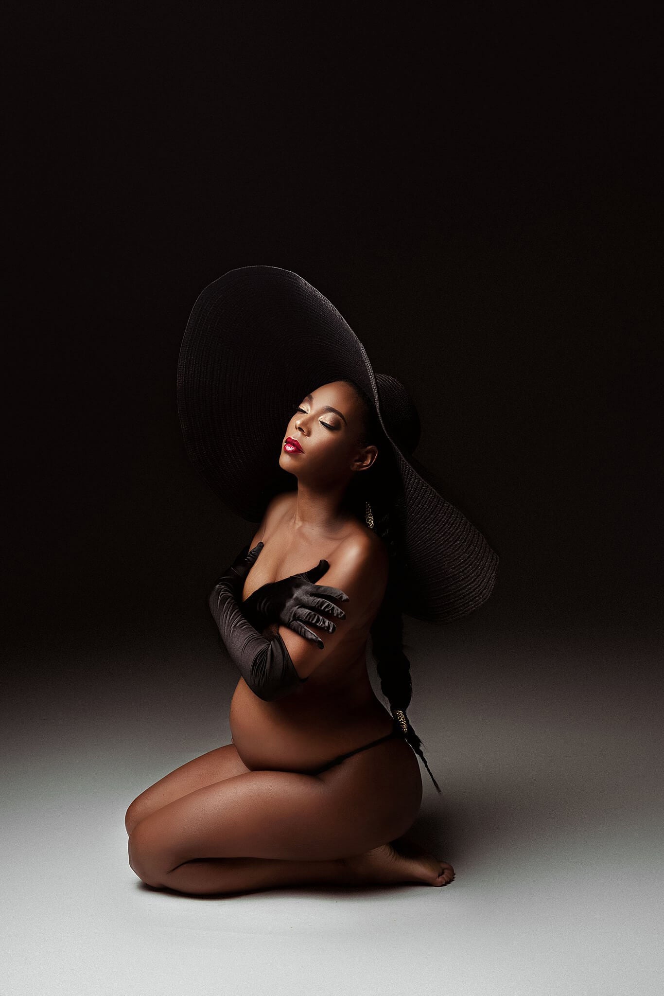 The woman is pregnant and sitting down on her knees. Her face is facing upwards and she has her eyes closed. She has long black gloves on. She is also wearing the product of the photo which is the big black hat. The hat is a real statement piece to use in your photography studio.