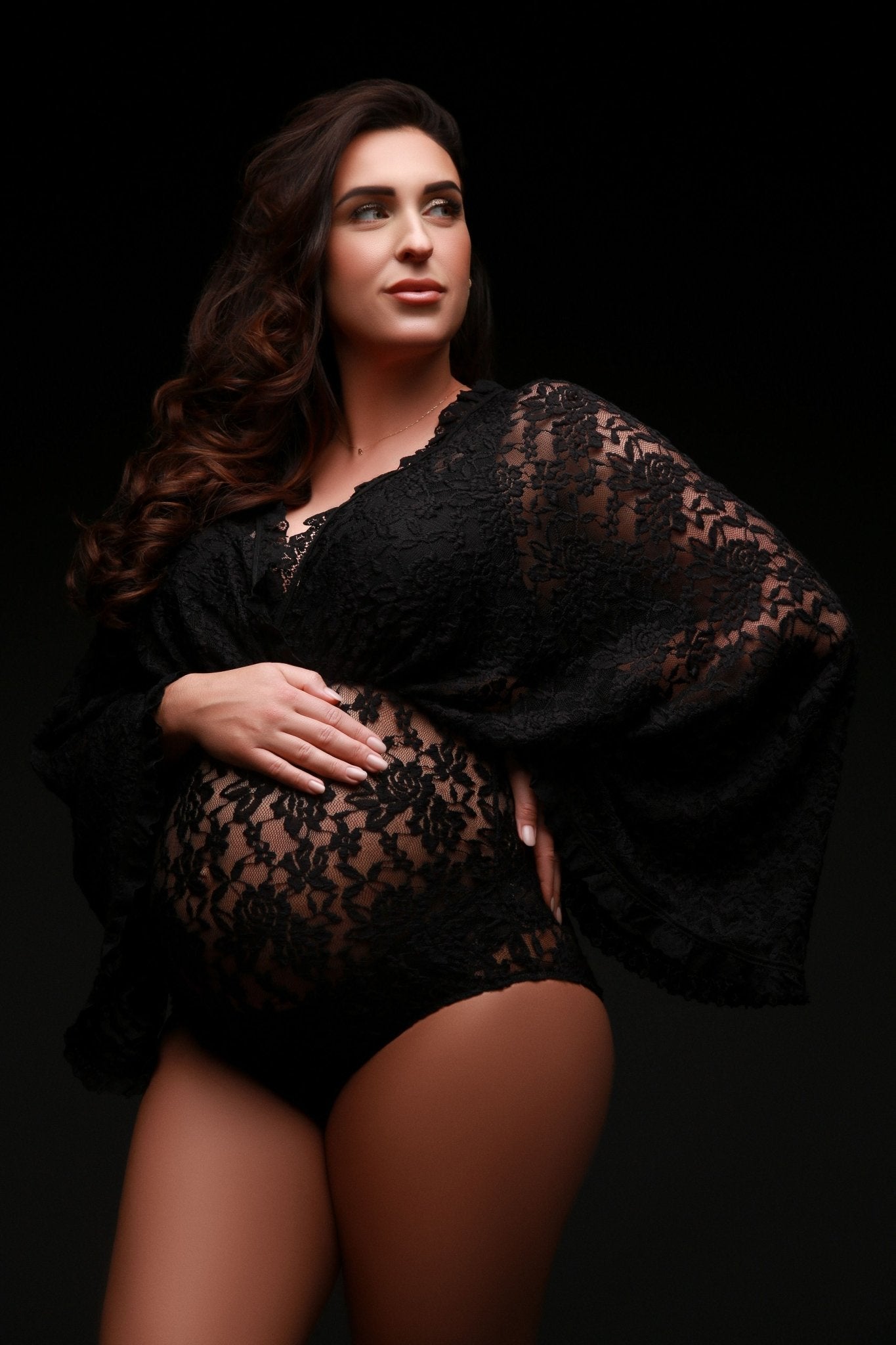 A pregnant woman is wearing a lace bodysuit. She is looking away from the camera. She has long brown hair. The bodysuit has loose sleeves and a v-neckline.