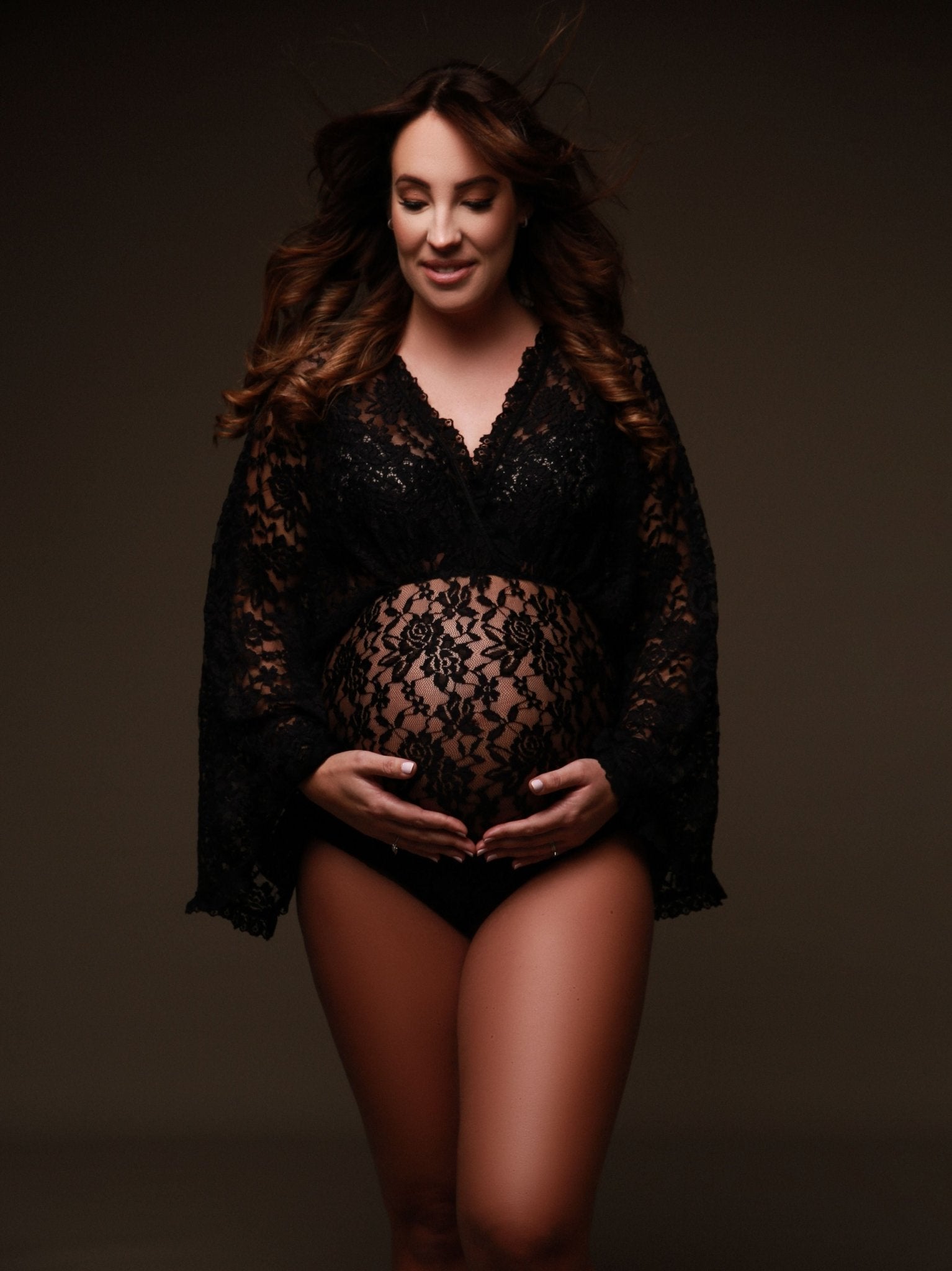 The model is pregnant and wearing a lace bodysuit. The top of the bodysuit is in a boho style. The woman has two hands underneath her belly. She has a soft smile on her face. The bodysuit is transparent.