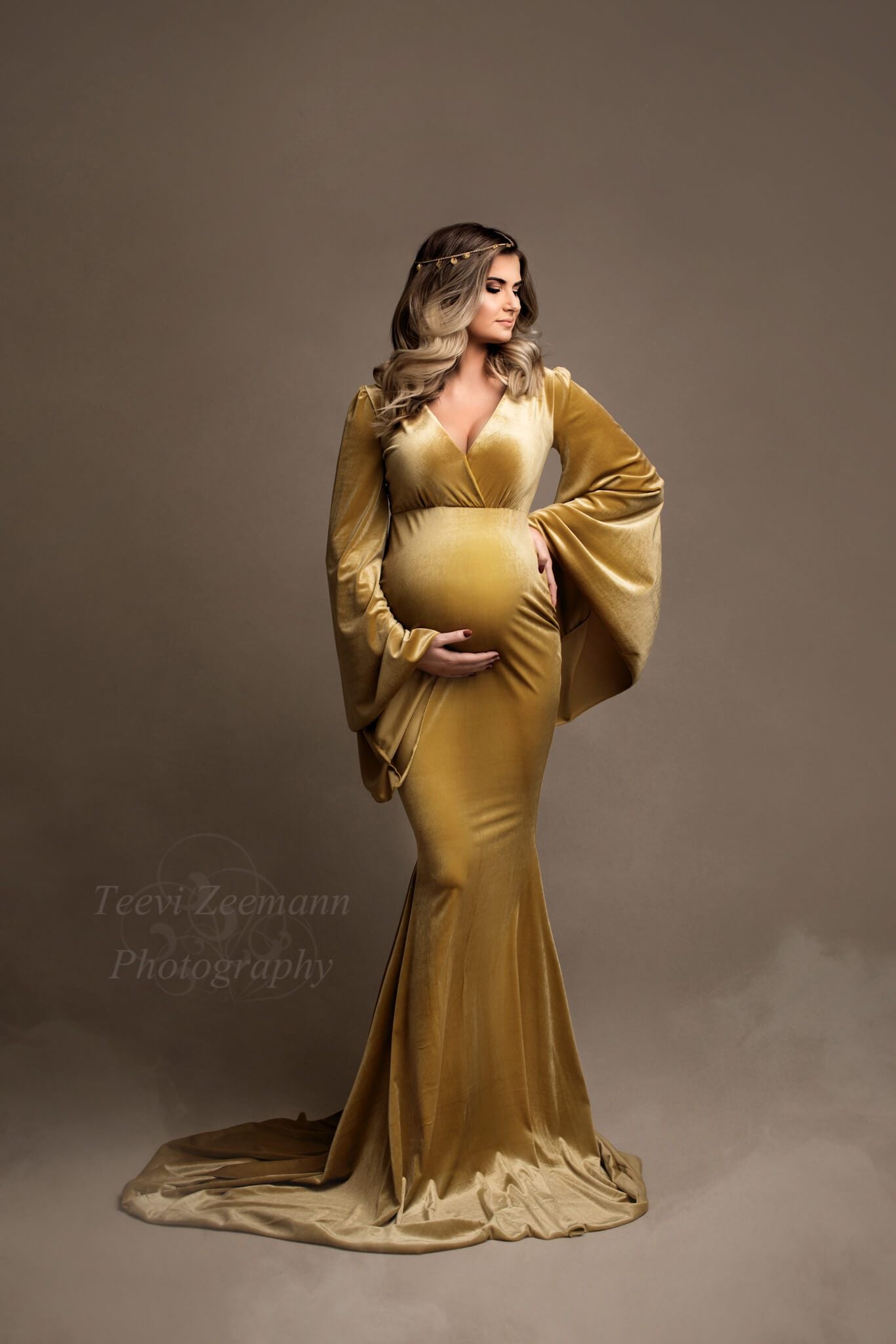 This model is standing in a studio with fake smoke around her. She is wearing a tight vevlet dress in the colour yellow amber. The sleeves are wide and flowy