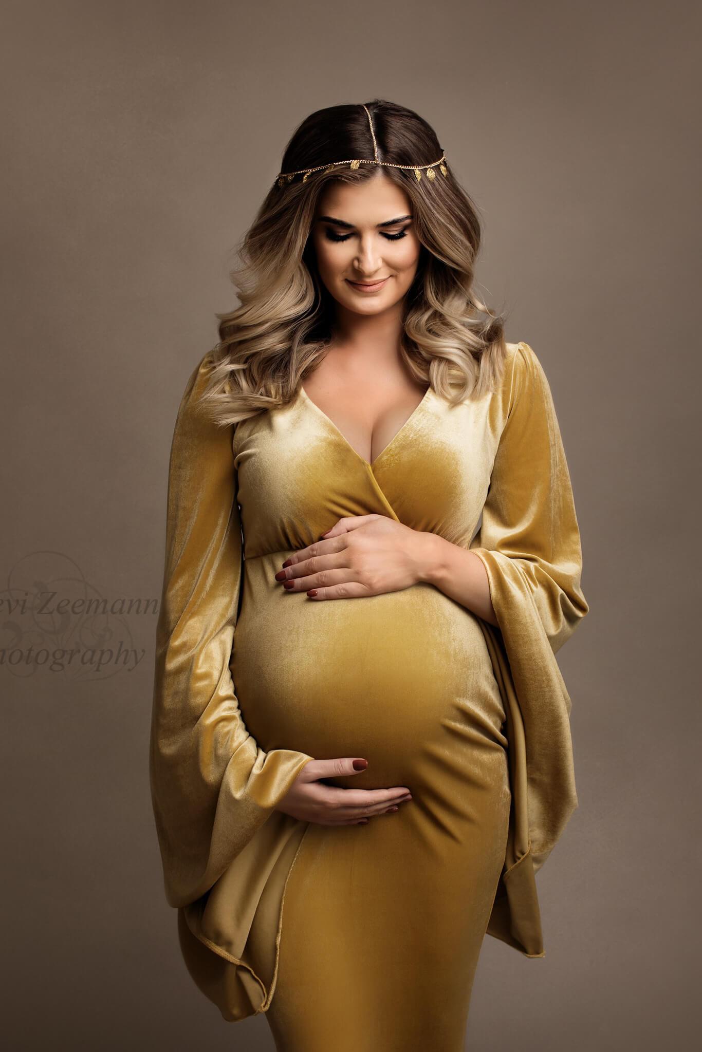 A close up photo of a woman that is pregnant. She is holding her belly and is wearing a yellow amber dress with a sweetheart neckline.