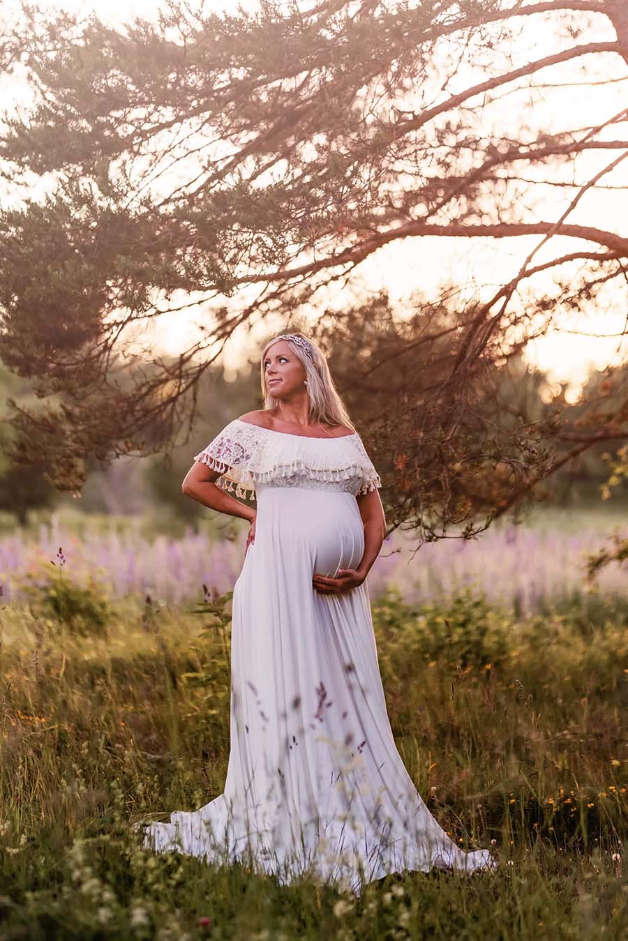 blond pregnant woman poses outside with a very green background wearing a dress made of jersey and lace in a white color. the top is off the shoulder with ruffle top and a jersey circle skirt.
