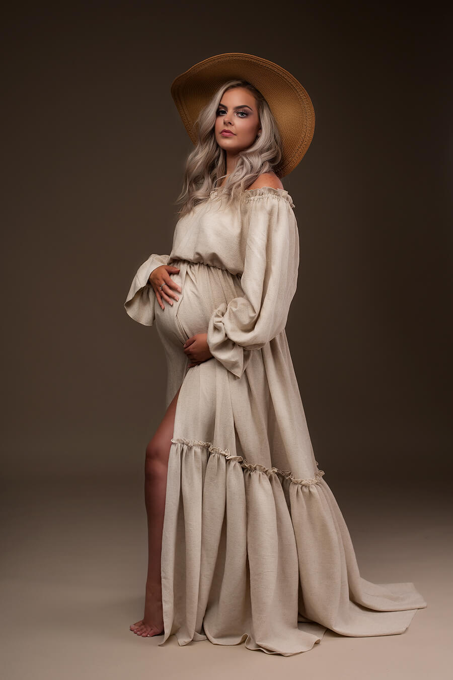 Blond pregnant model poses wearing a boho style maternity dress in sand color. She has a accessorizing hat to complete the look. She poses on her side, holding her bump and staring at the camera.