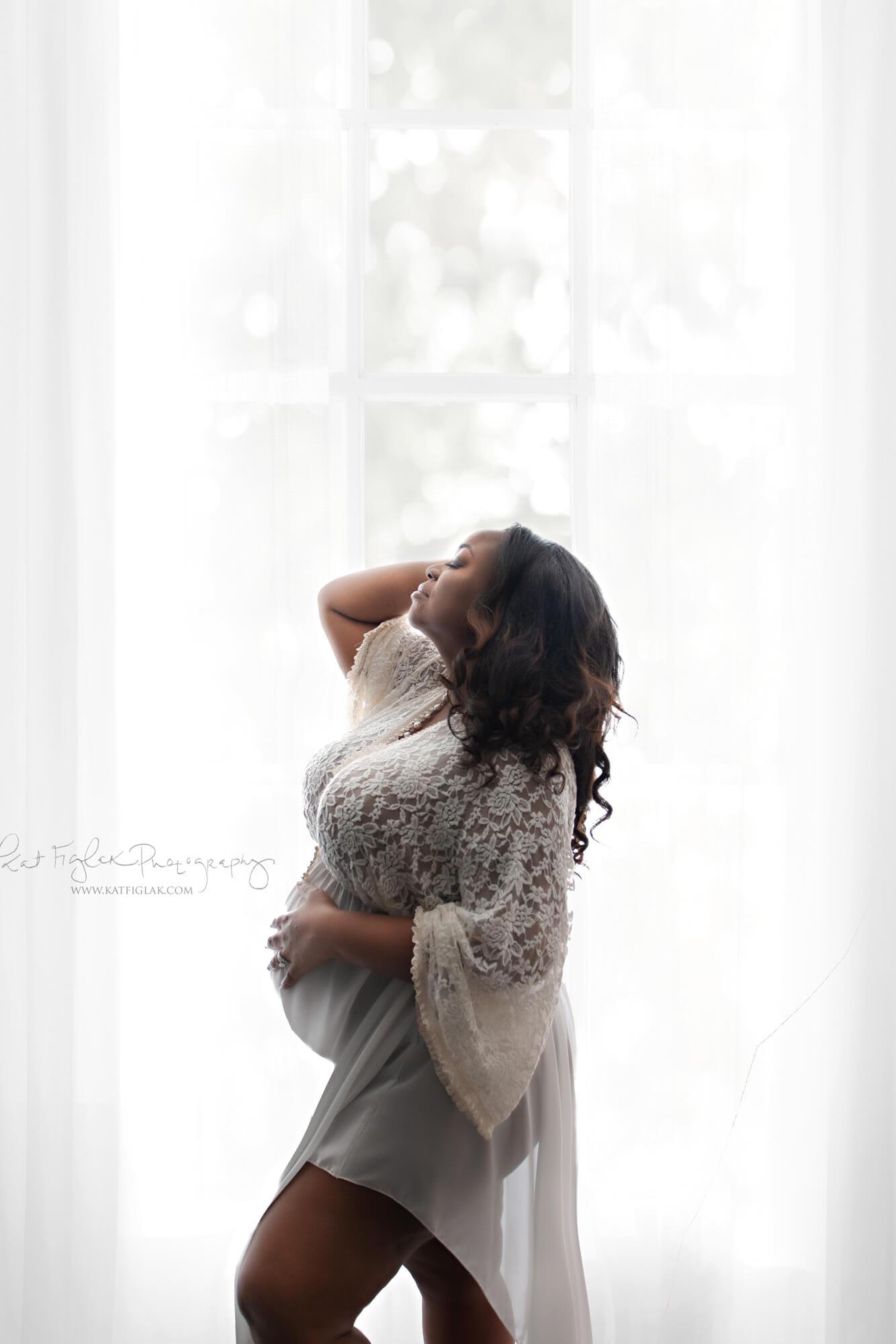 Pregnant model poses in studio during a maternity photosession. She holds her head with one hand and her belly with the other. She wears an off white long dress made of lace and chiffon. Her eyes are closed and facing up.