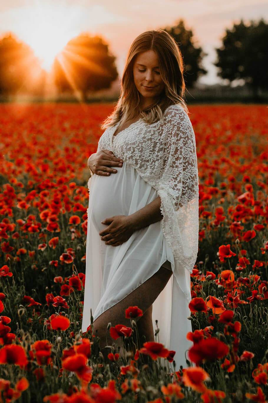 Blond pregnant model poses outside in a flower field wearing an off white long dress made of lace and chiffon. She has her eyes partially closed and looks down while holding her bump with both hands.