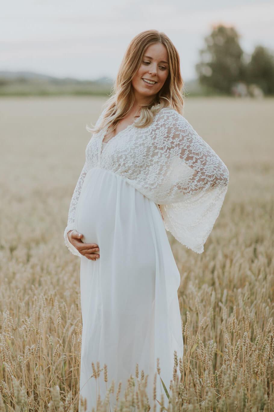 Pregnant blond woman poses outside wearing an off white long dress made of lace and chiffon. She stares to the side and holds her belly with one of her hands.