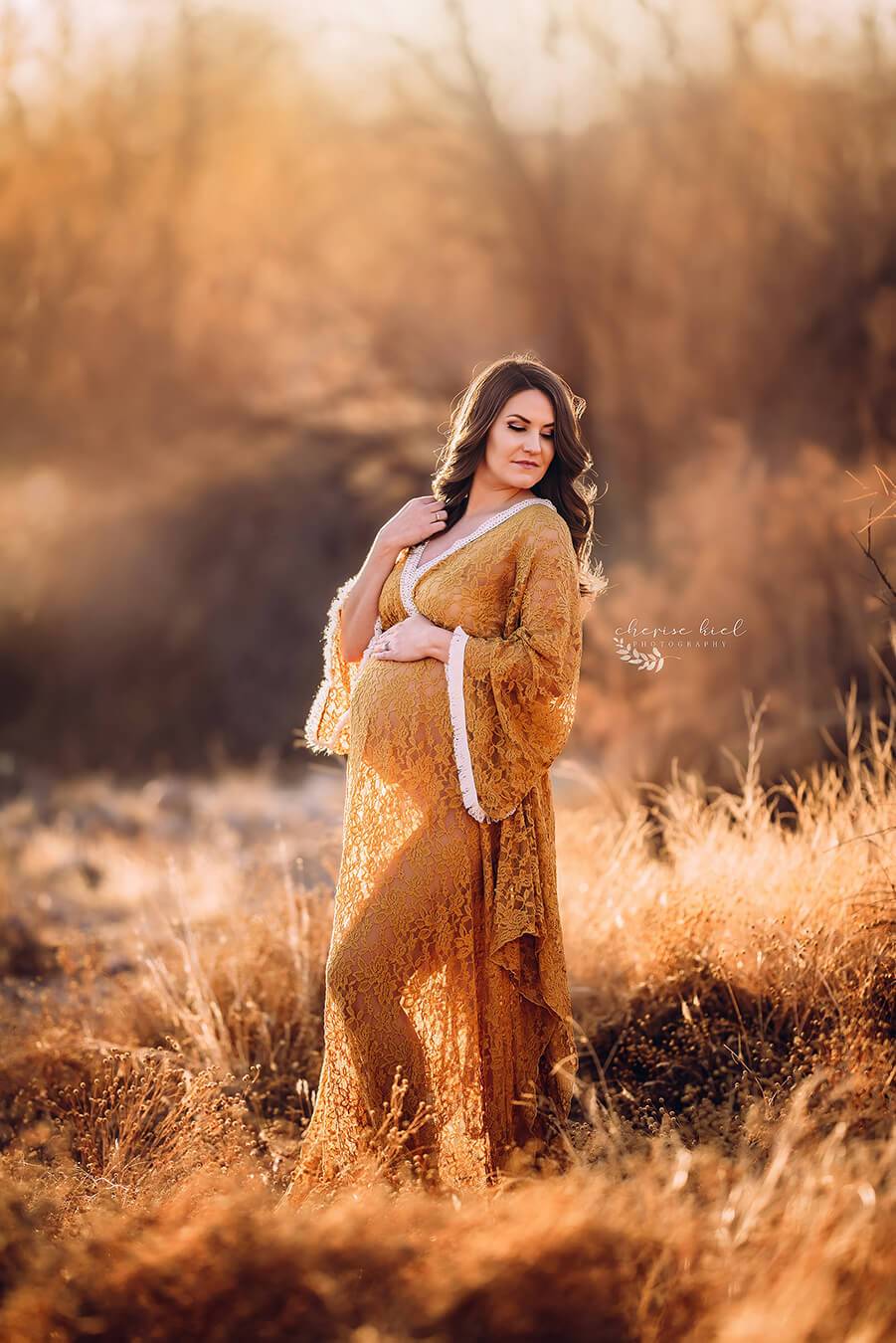 Pregnant model poses outside during a photo shoot. She wears a lace cognac dress, long enough to cover her feet. The dress has a low v cut neckline top and kaftan long sleeves. Her eyes are closed and she is touching her hair with one hand and her bump with the other one.