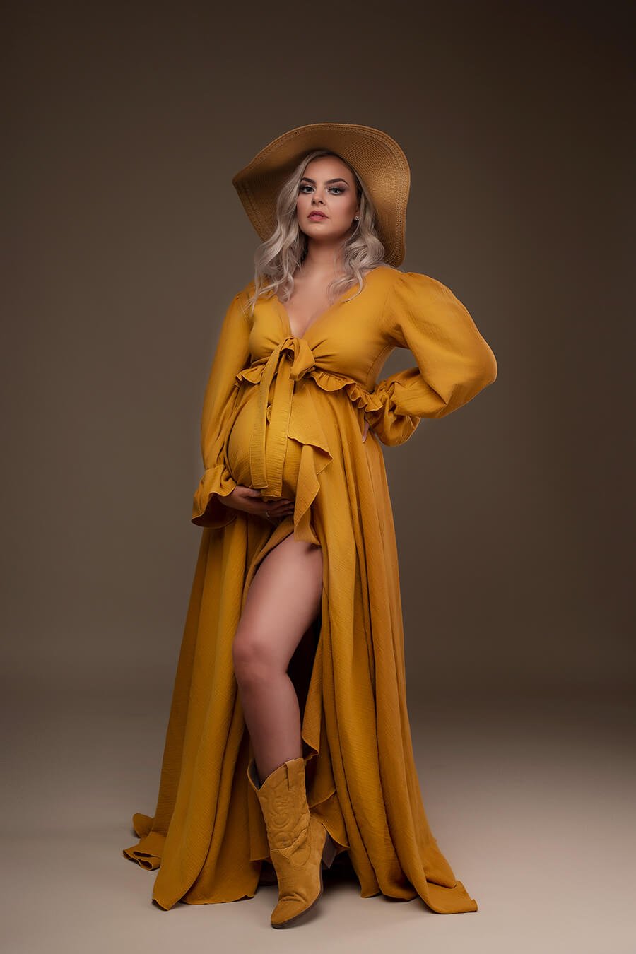 Blond model poses during a maternity photoshoot in a boho style. She wears an ocre long dress with ruffle details. The look is accessorized with a hat and boots.