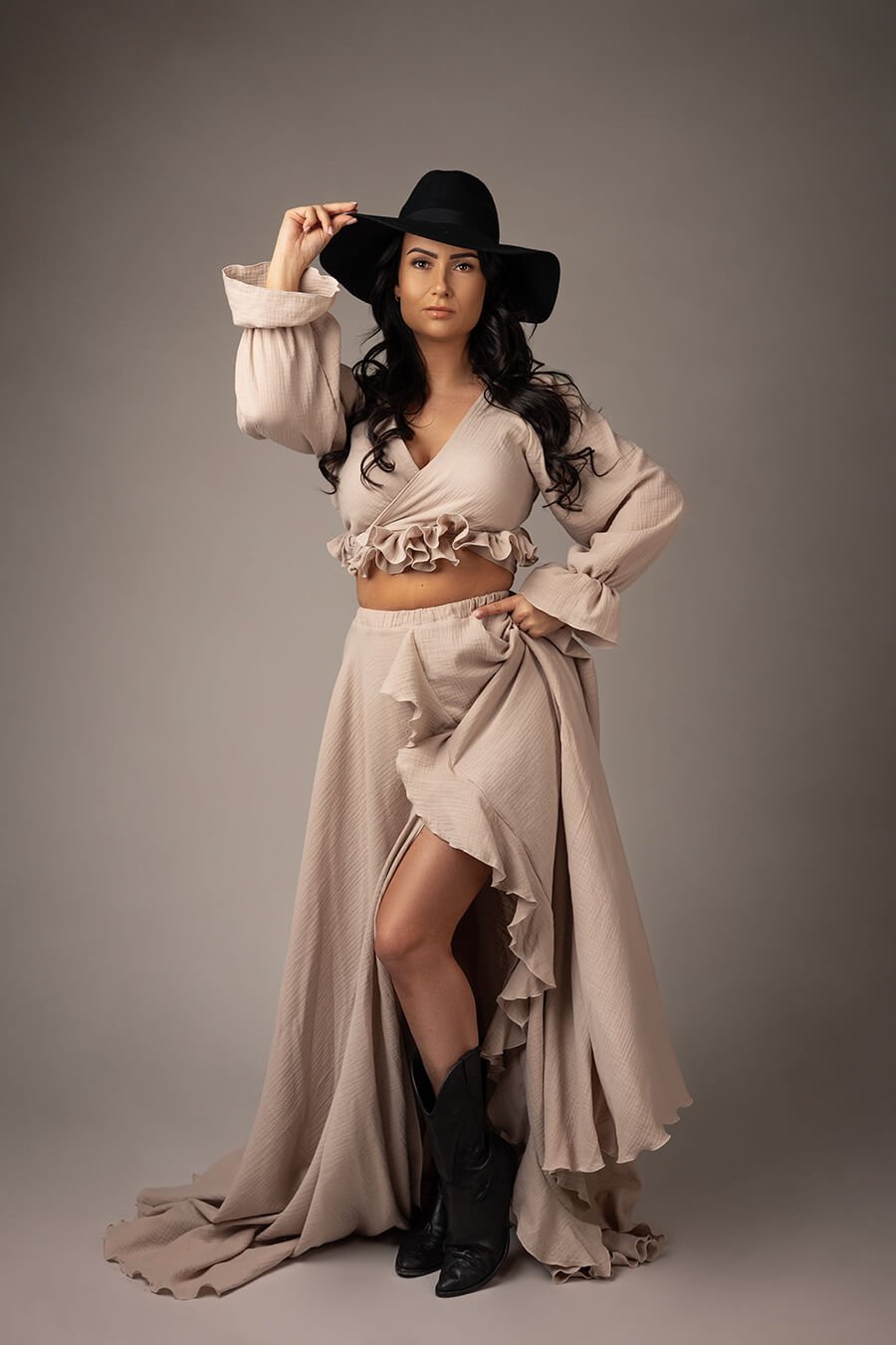 Brunette model poses in a studio wearing a sand set in boho chic style. she has a black hat and black boots to match the style. she is wearing a long skirt with ruffle details and a short top with long sleeves, also with ruffle details.