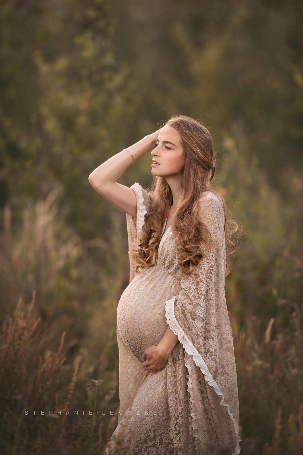 Blond pregnant model poses outside wearing a lace dress. The close up is made with her profile while she looks away, holding her bump with one hand and touching her hair with the other one.
