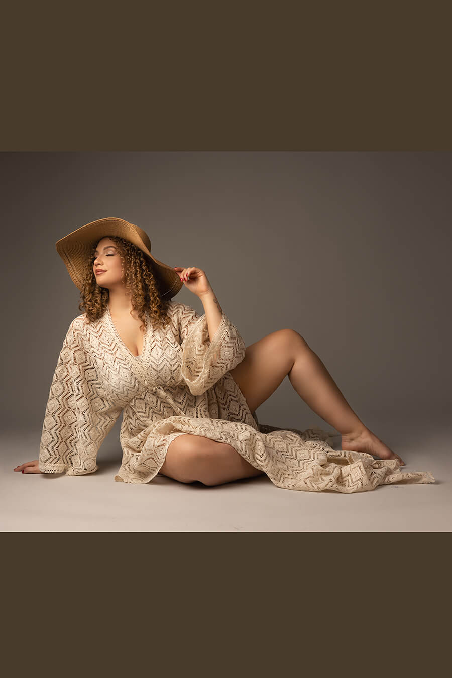 model poses sitting on the floor of a studio during a boho chic photoshoot. she has one hand on the floor and the other one touching the hat. her legs can be seen through the asymmetrical skirt.