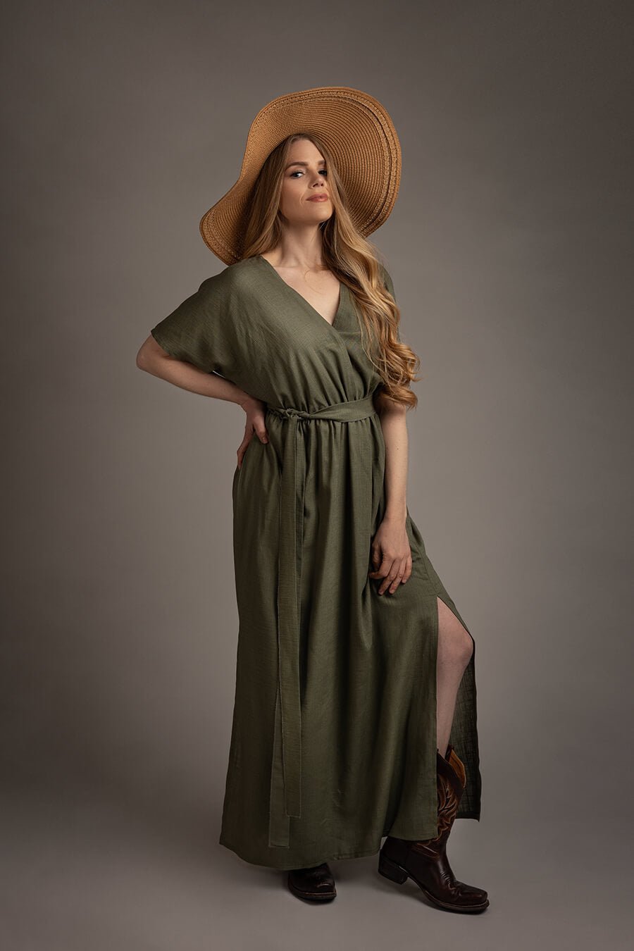 blond model poses in a studio wearing a boho style dress in green color with short sleeves and a split on the skirt. the top features a low v cut neckline. she has a brown hat to match the western boho chic style.