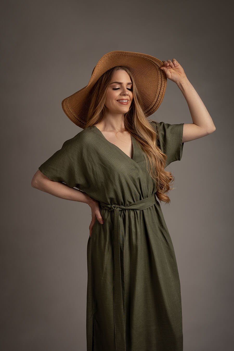 blond model poses in a studio wearing a boho style dress in green color with short sleeves and a split on the skirt. the top features a low v cut neckline. she has a brown hat to match the western boho chic style.