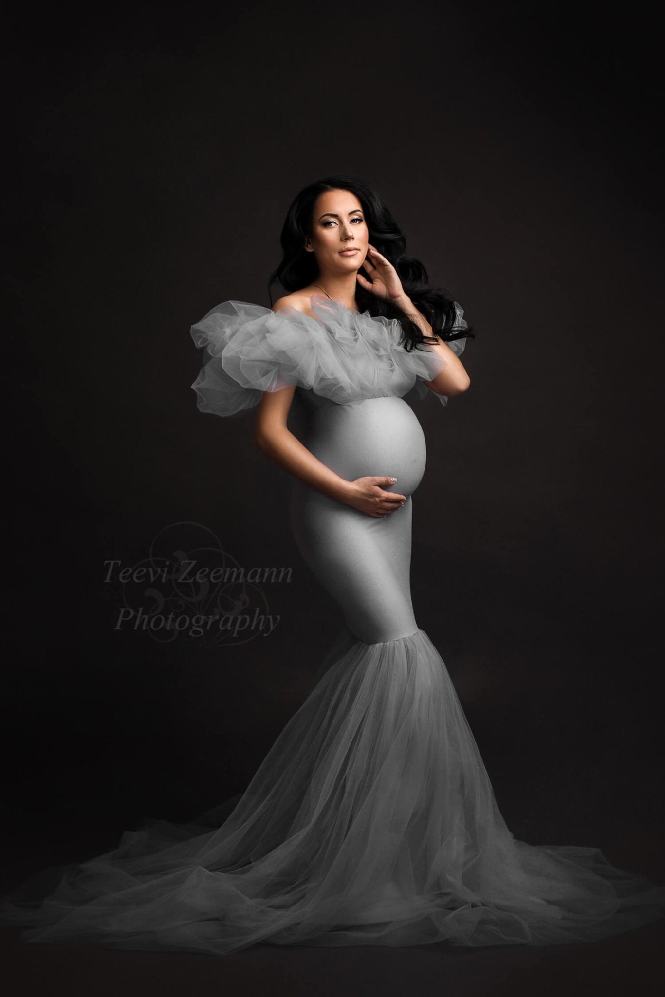 The model in this photo is wearing a long grey dress. The dress has a unique effect because it is made out of tulle and chiffon. The dress is for maternity woman. It has a off shoulder model and a mermaid skirt.