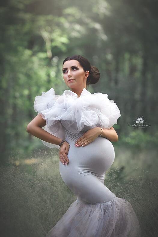 This pregnant model is standing outside in the woods. She has a bun in her hair. She is wearing a grey dress with tulle at the top. The dress has a extra effect.