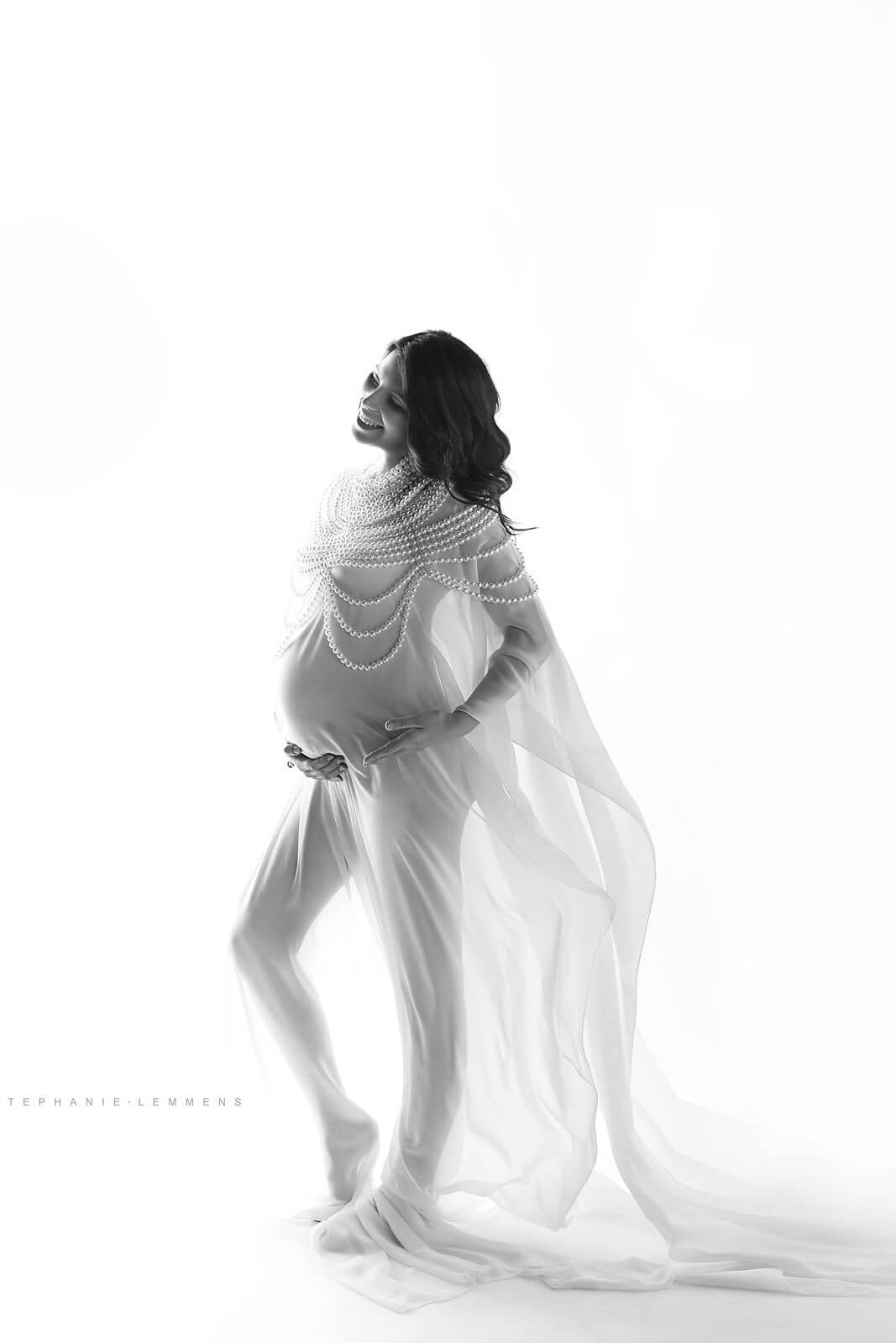 brunette pregnant model wearing a white and long chiffon cape. above the cape she has a very big pearl necklace to match the style.