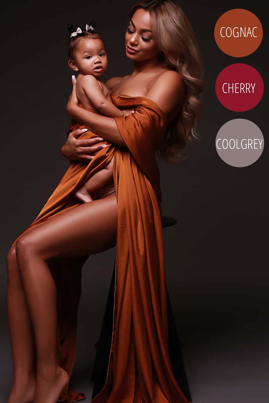Model poses together with her baby daughter for a studio photo shoot. She holds her baby wrapped in a silky cognac draping fabric. She looks to her baby and her baby faces the camera.