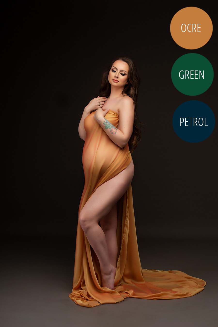 Pregnant model poses in a dark studio wearing a draping fabric in ocre color. She holds the fabric against her chest and has her belly covered with the draping piece. She looks to the front with her eyes semi open.