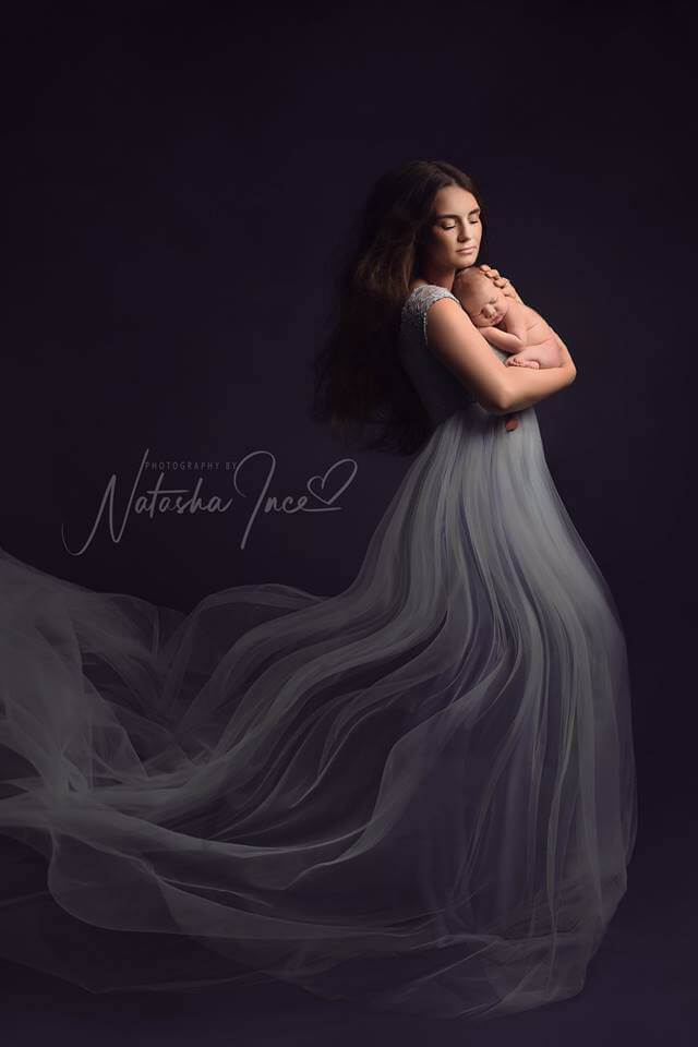 model poses holding her newborn. she has her eyes closed and wears a tulle dress with a lace top. the dress is light blue.