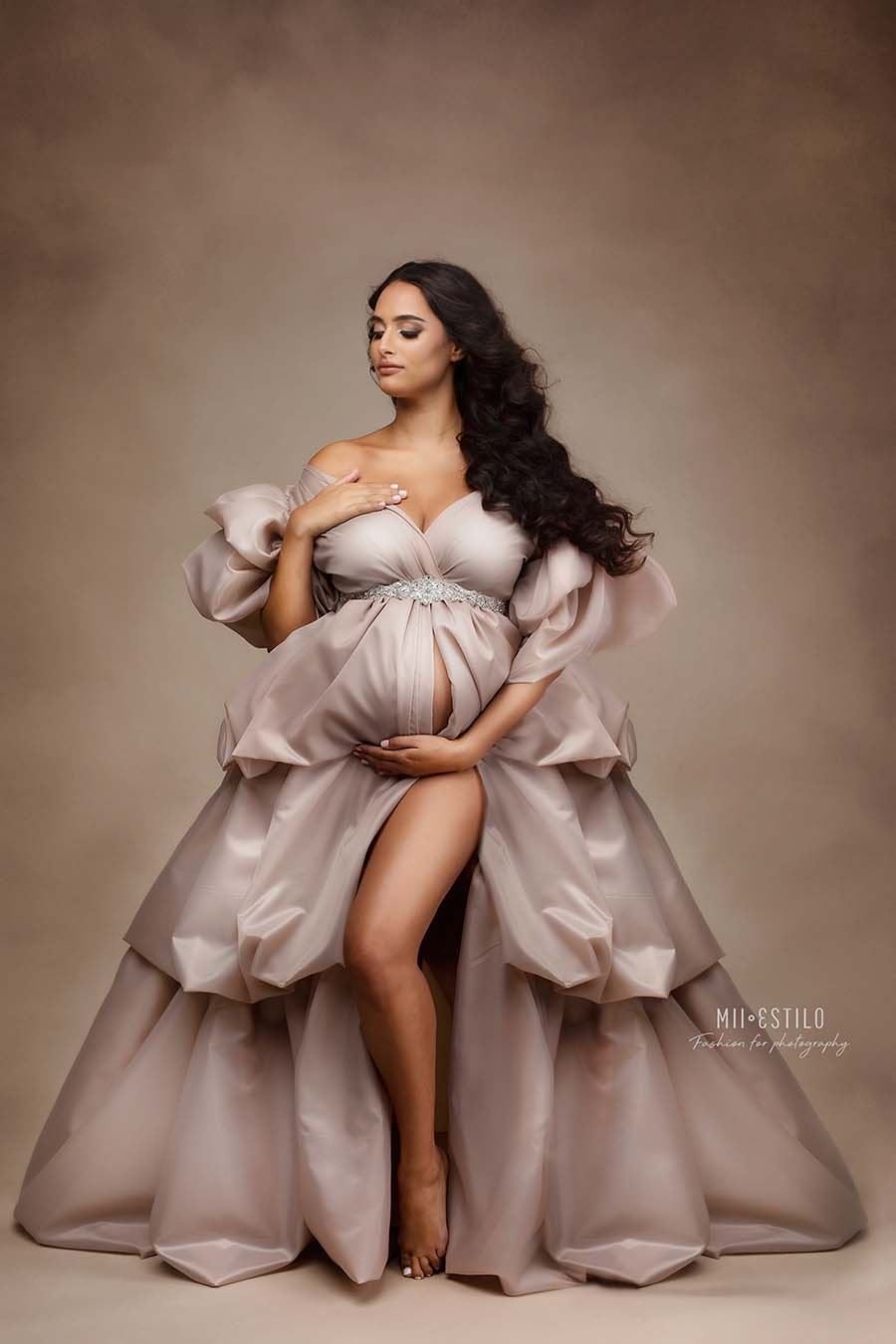 brunette pregnant woman poses in a brown studio wearing a long and big dress made of taffeta in sand color. the dress has several layers to create more volume.