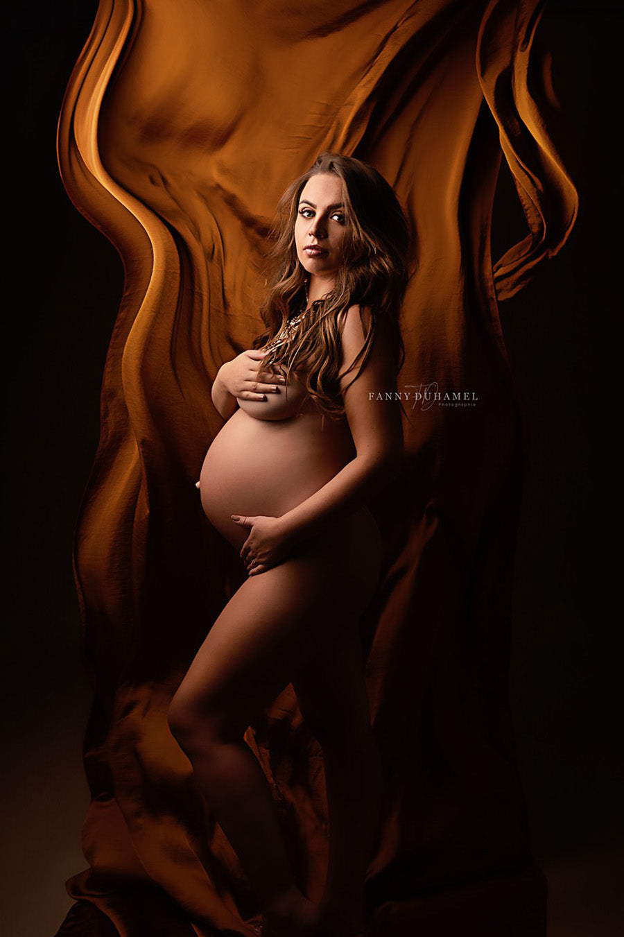 brunette model poses in a dark studio in front of a silky fabric in cognac color. the fabric has been thrown before the close up. the model covers her breast.