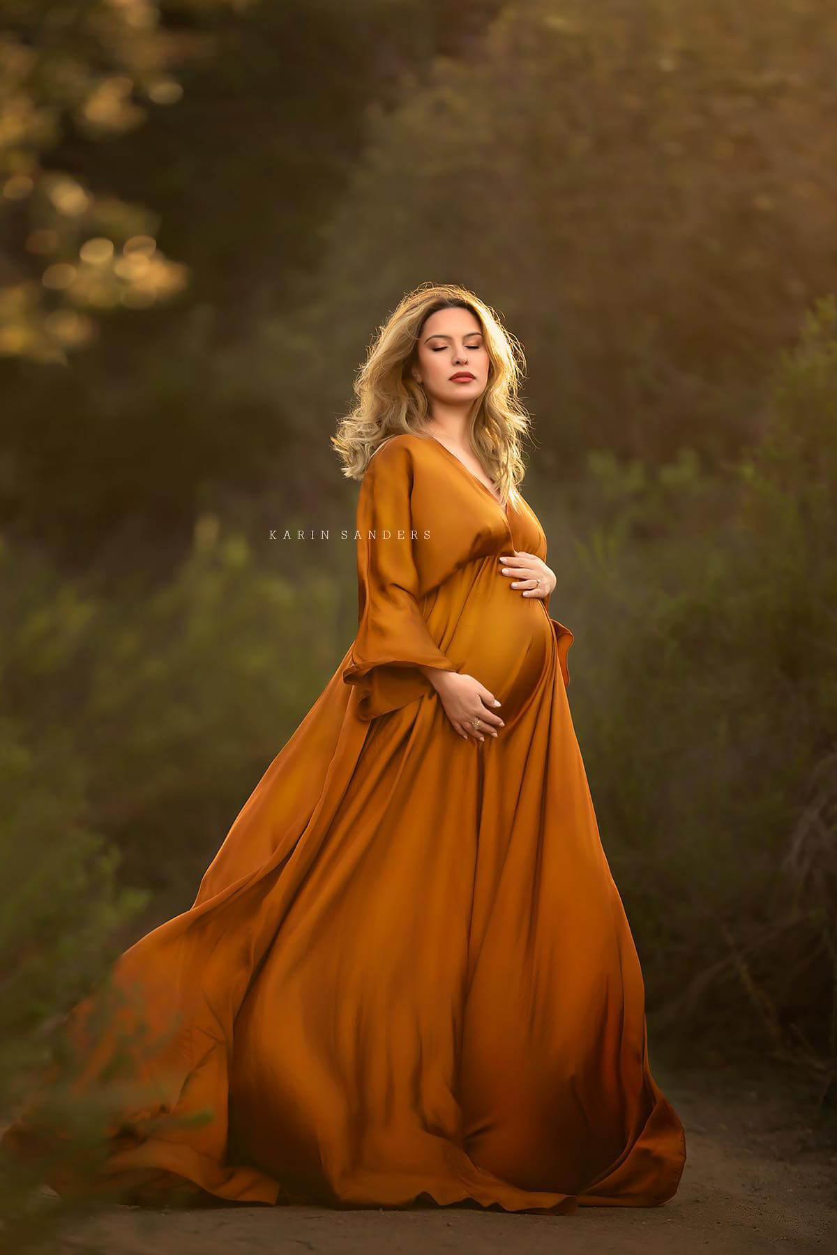 Blond pregnant woman poses outside wearing a long cognac silky dress with kaftan sleeves and a low v cut neckline. She has her eyes closed and holds her bump with both hands.