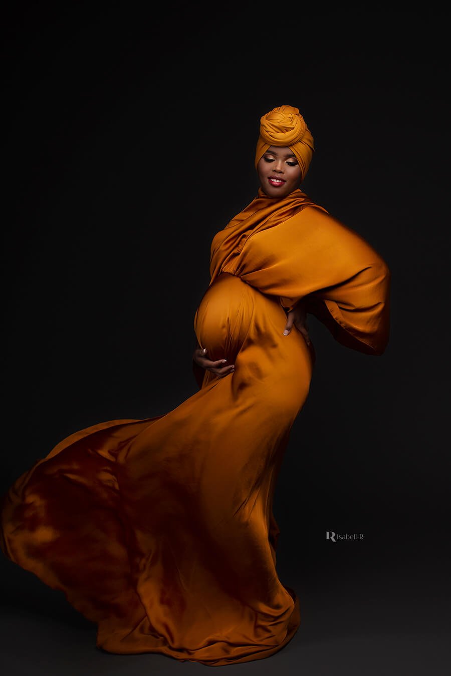 Pregnant model poses in a studio wearing a cognac long dress made of silk. She poses on her side and has her eyes closed. one of her hands is holding her back and the other under her bump. She has a matching piece of fabric on her head.
