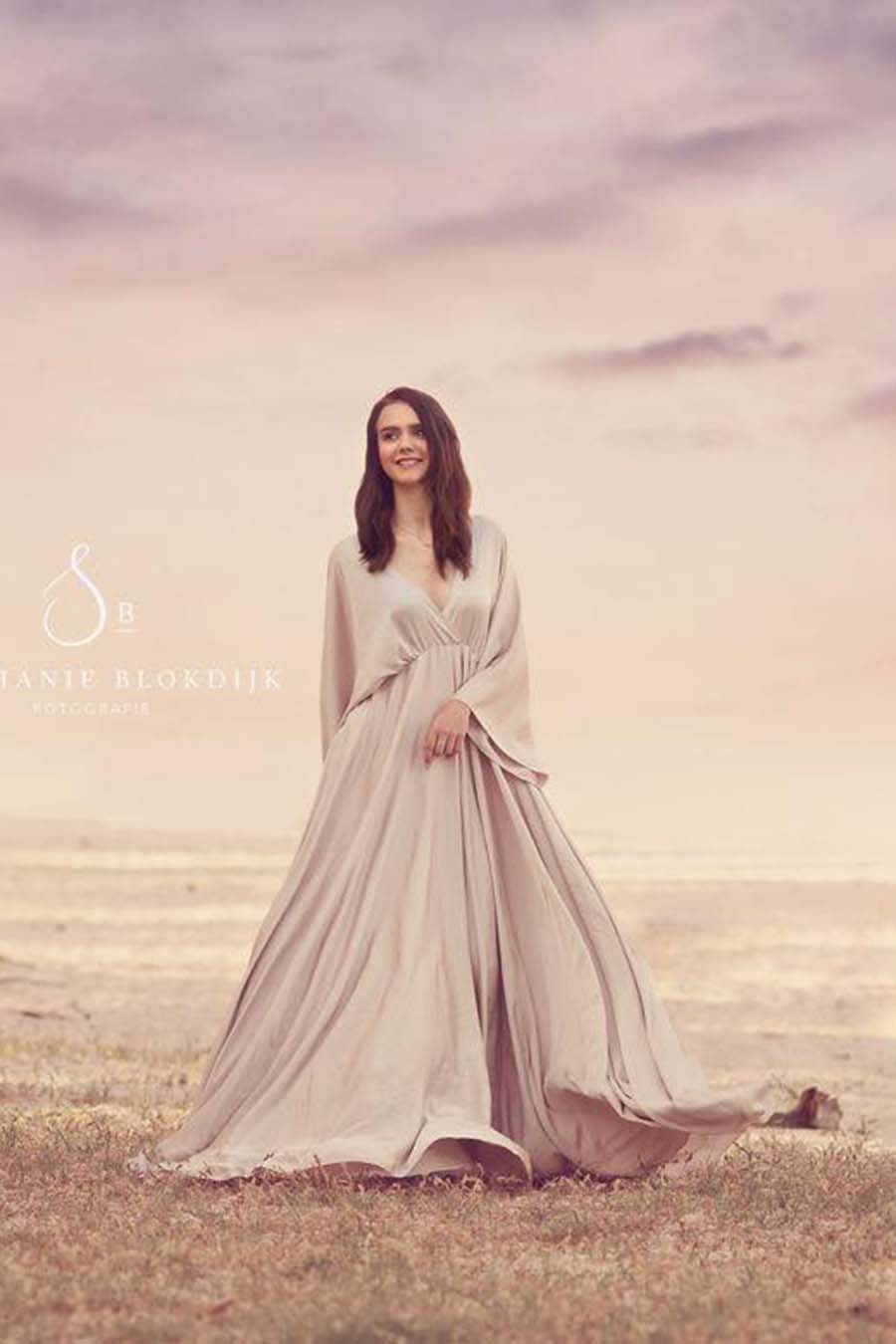 Brunette model poses outside with a mii-estilo dress made of silk. She looks to the side with her eyes open and has one of her hands in the front of the dress. The top features a low-v cut neckline. The skirt is wide and long and the dress is made in a sand color.