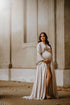 Brunette model poses outside with a mii-estilo dress made of silk. During this maternity photo session the model shows one of her legs through the split on the skirt. The dress features a low-v cut neckline and kaftan sleeves. The model holds her bump with both hands and looks to the side.