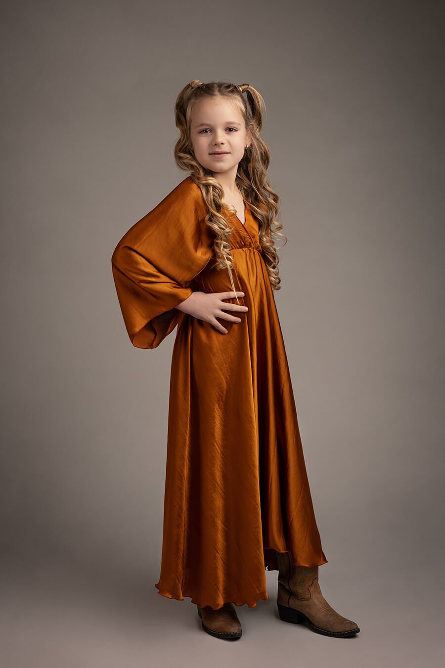 blond girl poses in a studio during a boho chic photoshoot. she is wearing a long silky cognac dress in a boho chic style and wears boots to match the look.