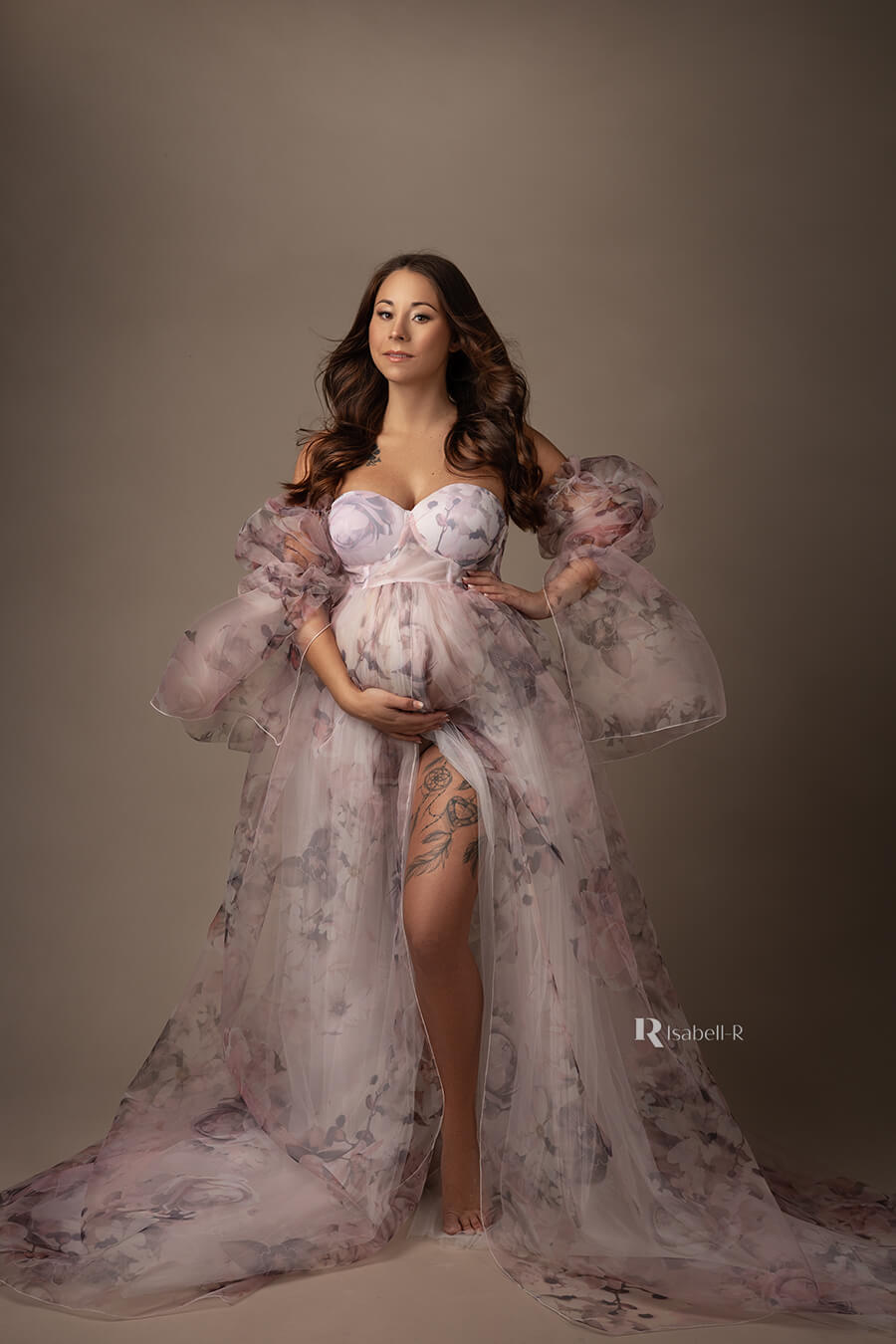 brunette model poses in a studio wearing a flower patterned dress in organza and tulle. the dress has wide attached sleeves made of organza and a split on the skirt where one of her legs can be seen.