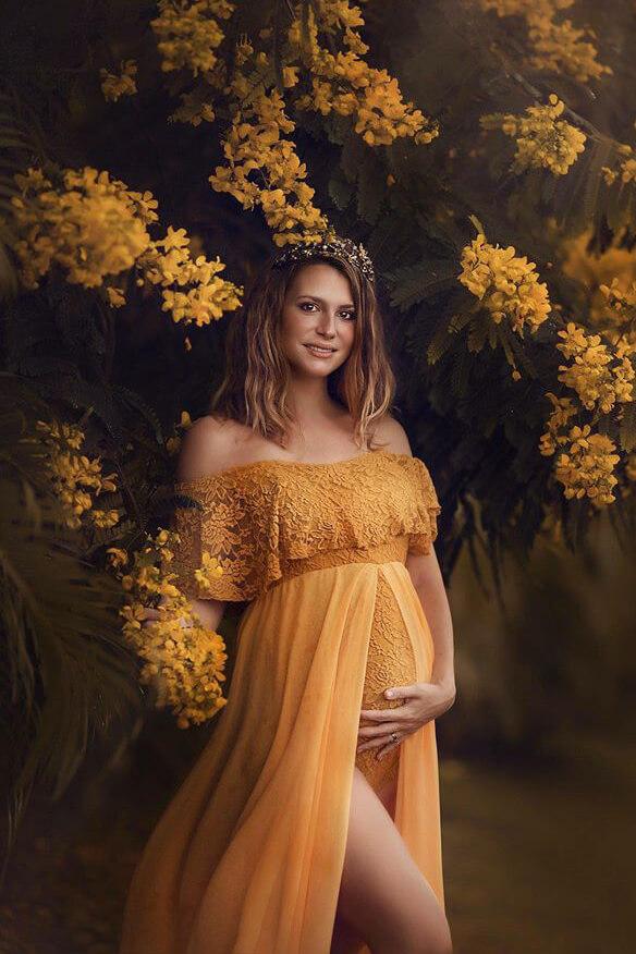pregnant woman poses next to a tree with yellow flowers while wearing a yellow maternity dress with a chiffon long train and a bodysuit underneath made of lace and jersey. the model has a flower crown and stares at the camera.