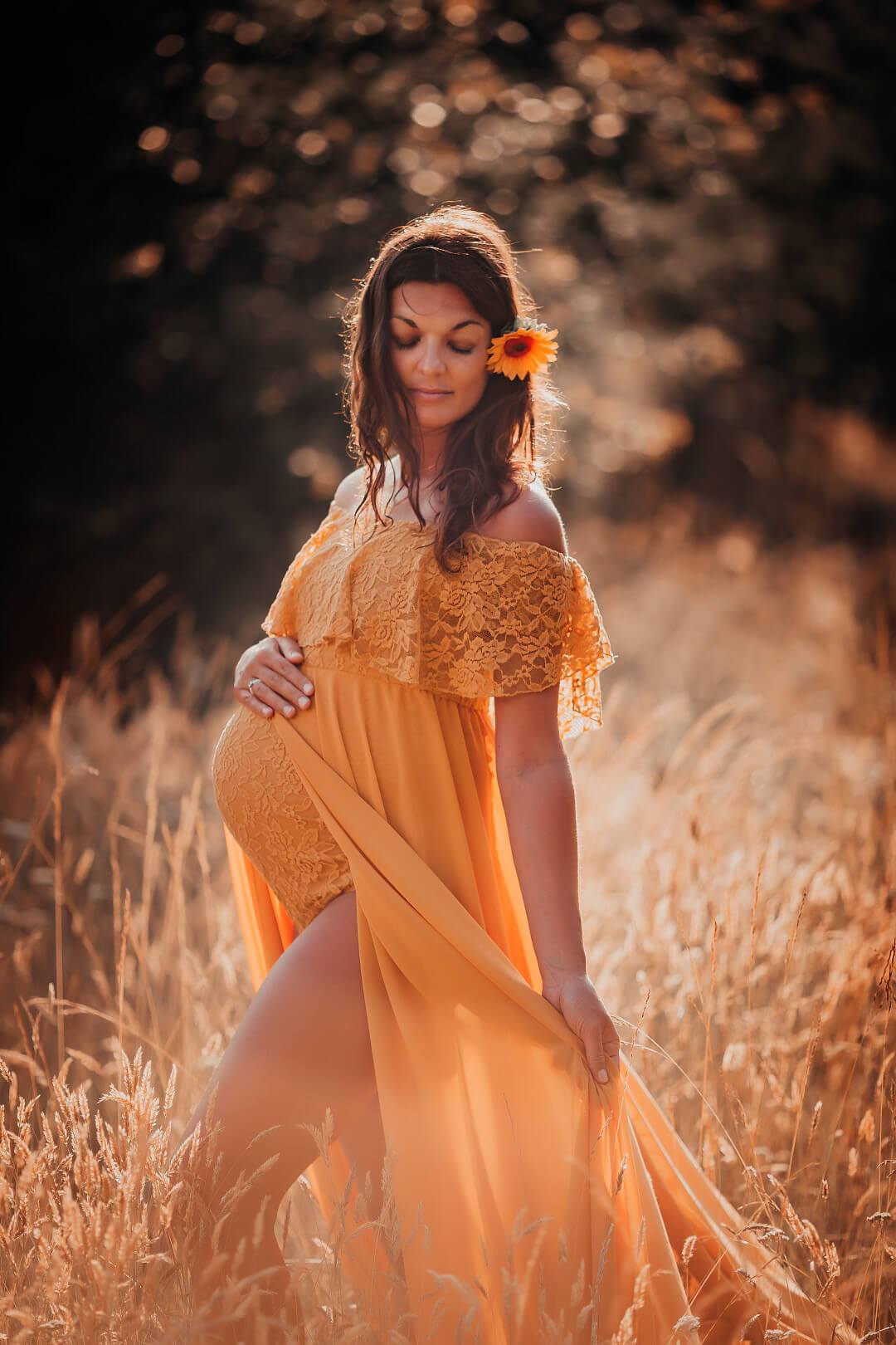 pregnant woman poses outside wearing a yellow maternity dress made of lace, jersey and chiffon. she has a sunflower on her head.