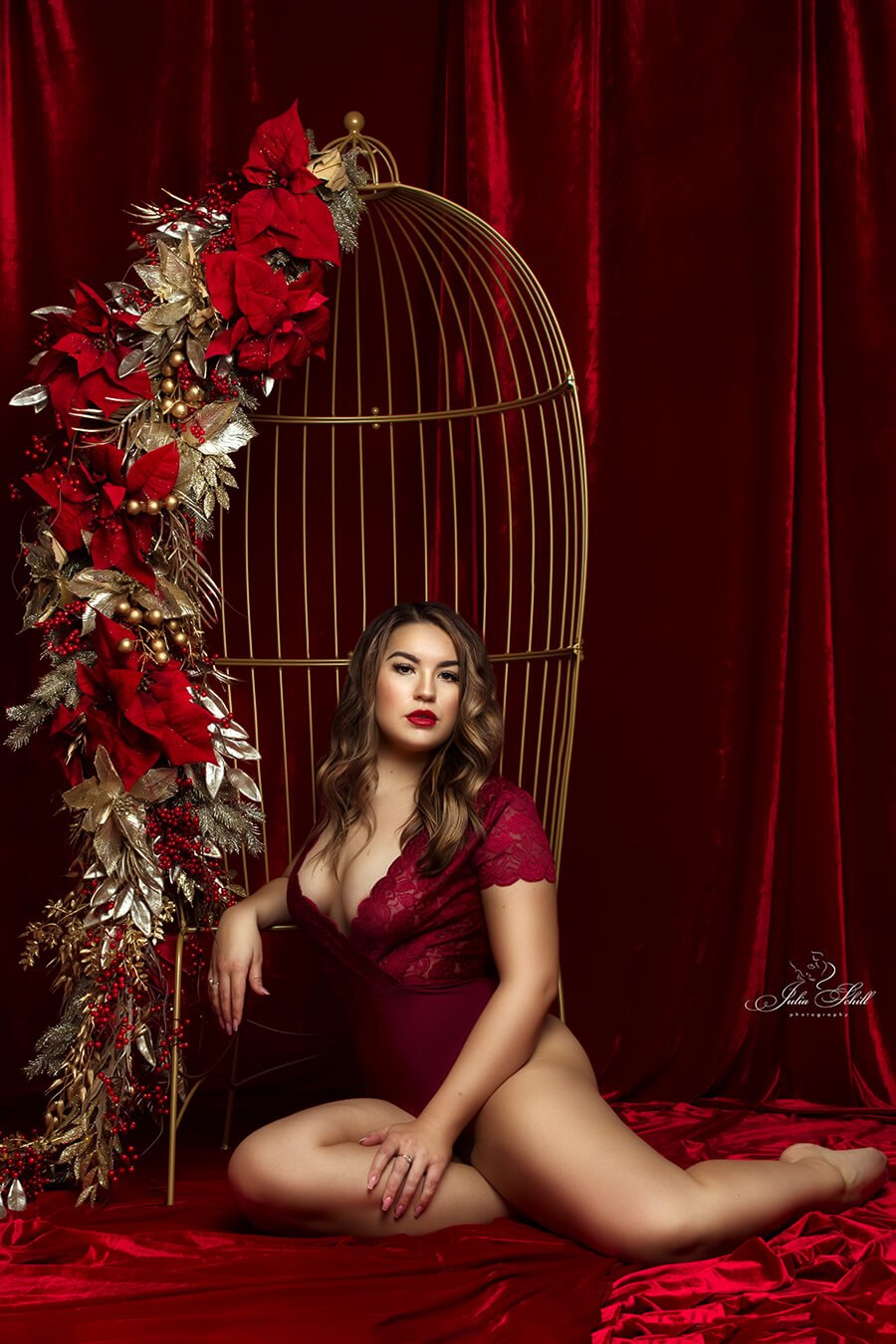 dark blond model poses in a studio, sitting next to a metal chair that resembles a cage with several red details. she is wearing a red bodysuit during a boudoir session.