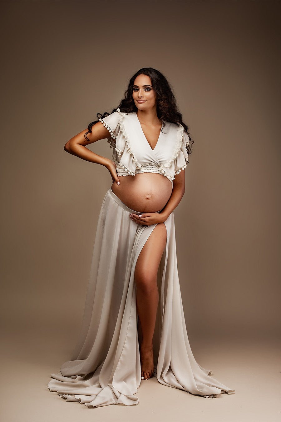 pregnant woman poses in a studio wearing a chiffon maternity set in sand color. her bely is uncovered and she is posing her hands gently in there. the top features a low v cut neckline with ruffled chiffon details.