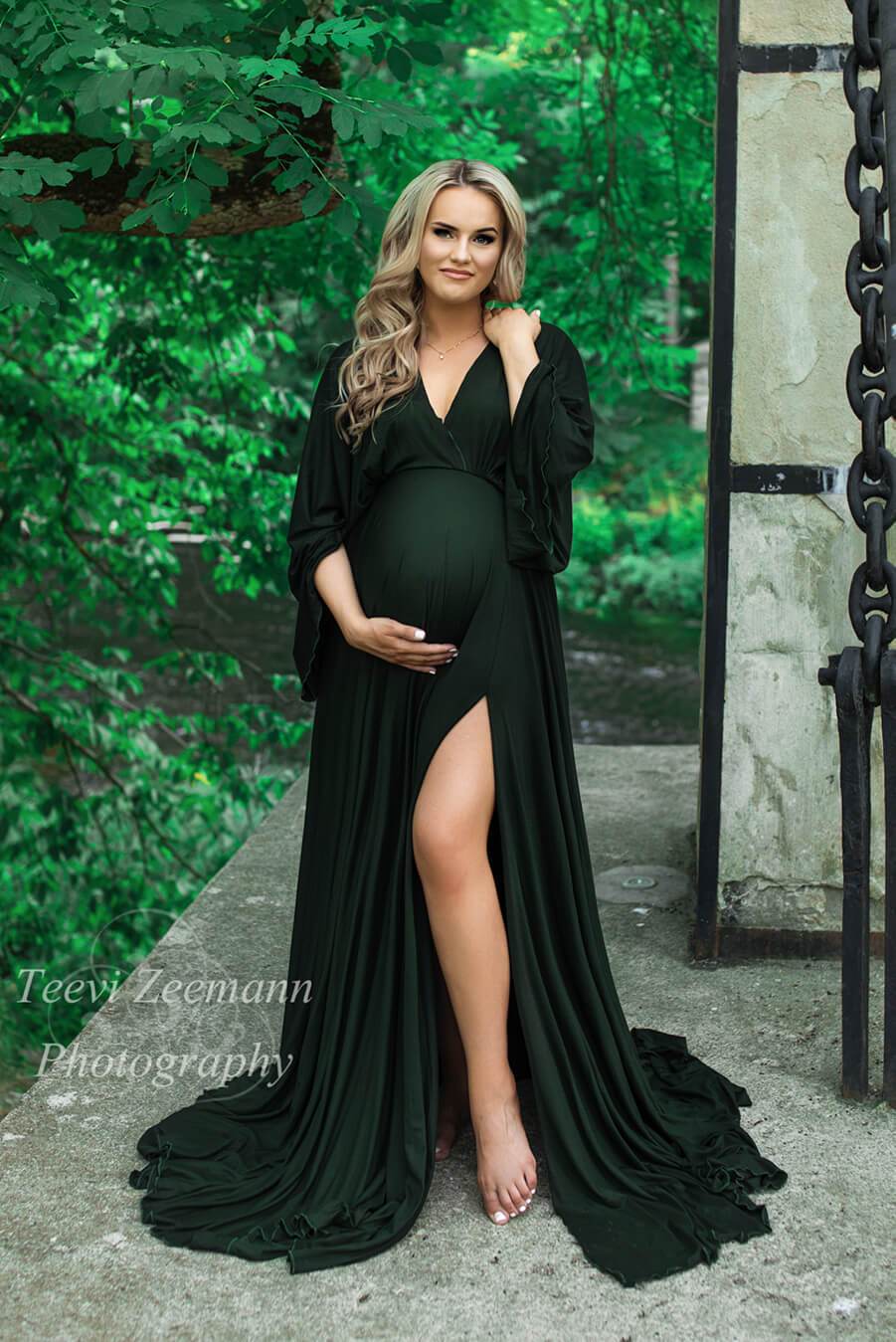 A pregnant woman is standing in a garden. She is wearing a dark green dress with a split by the leg. The sleeves are boho styled