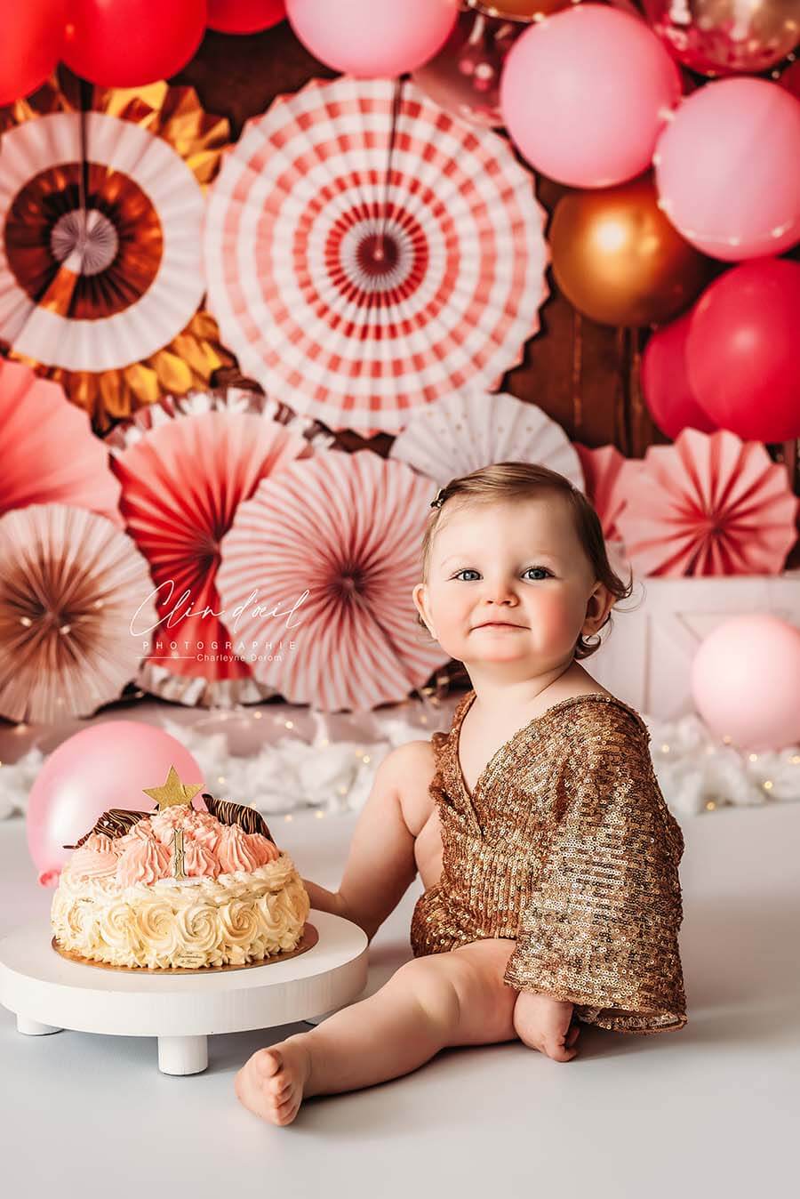 A babygirl is sitting by a birthday cake. There are balloons and other decorations behind her. She is wearing a little baby romper in the color gold. the romper is really sparkling.