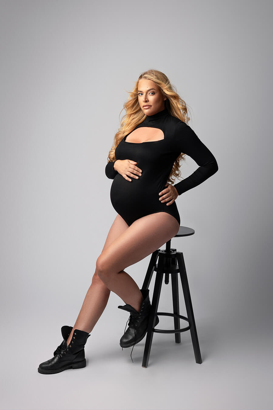 blond model poses sitting on a high chair and wearing a black jersey bodysuit with long sleeves, keyhole neck and a turtleneck neckline.