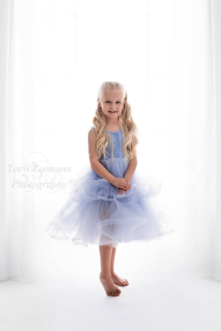 little blond girl poses wearing a lace and tulle light blue dress. her hands are in the front and she is smiling to the camera.