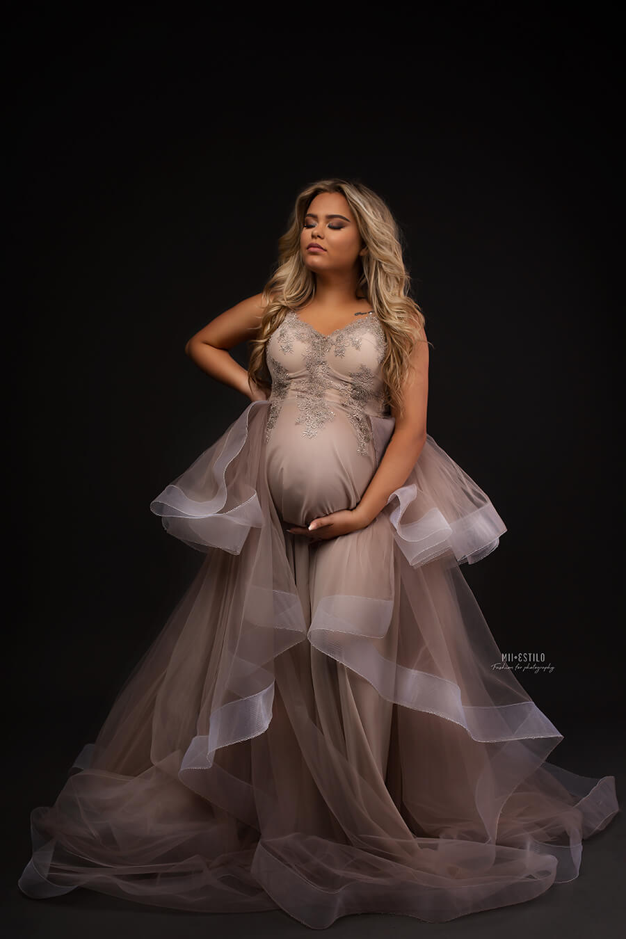Pregnant blond woman poses in a studio wearing a dress made of tulle and bridal 3d lace. She has her eyes closed and looks up while holding her bump with one of her hands. The dress has a sand color and the skirt is made in layers of tulle.