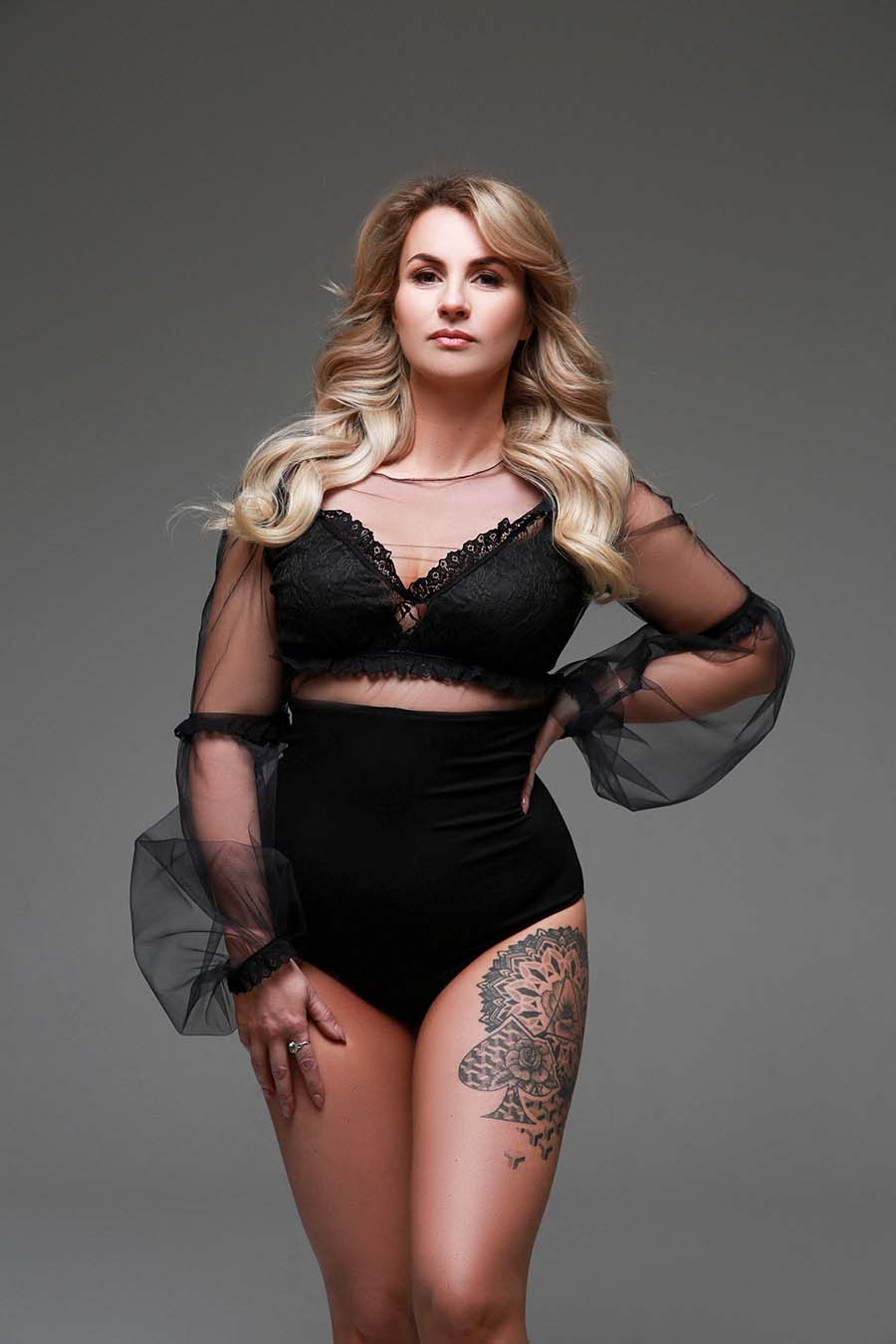 This woman is posing in the Madeline Bodysuit. The bodysuit has sheer sleeves and a lace top. The bottom half is black jersey.
