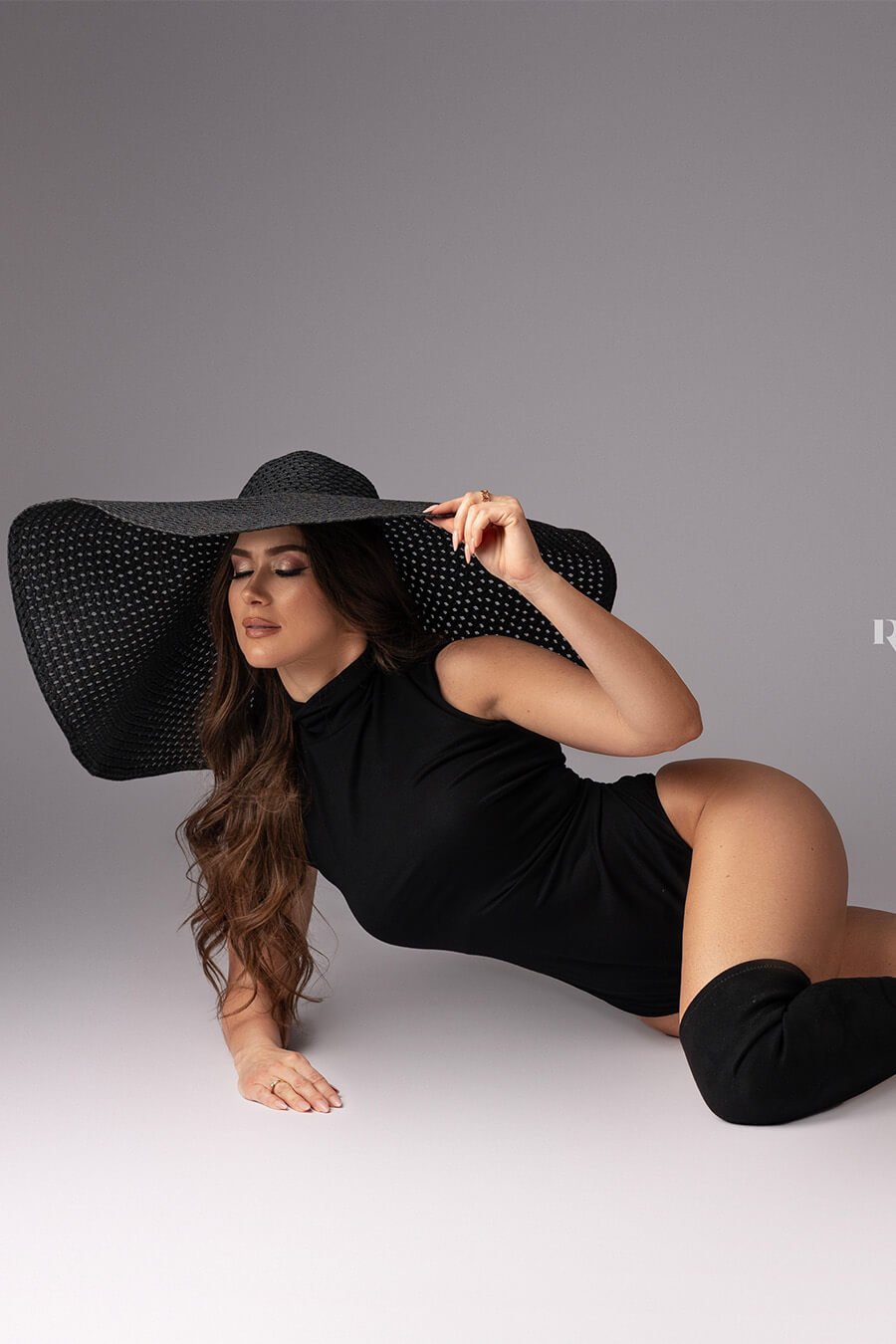 brunette model poses laying on the studio floor wearing a black bodysuit with a turtleneck and matching socks. she has a very large black hat to match the style. one arm leans on the floor and the other hand is holding a piece of the hat. her eyes are closed.