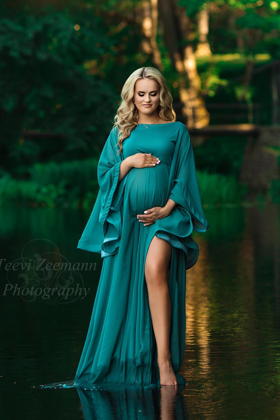 A woman is standing in a lake. She is wearing a long dress in the colour petrol. She has long blonde hair and looking down. The dress has a split by her left leg